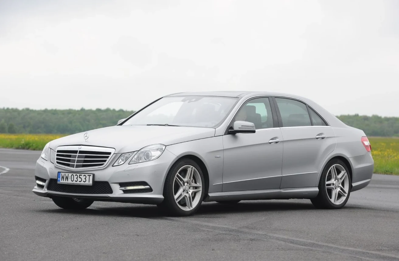 Używany Mercedes E-Klasa W212 - najlepsza limuzyna na rynku? srebrny sedan marki Mercedes-Benz zaparkowany na asfaltowej powierzchni, samochód ustawiony pod lekkim kątem z widocznym przodem i bokiem, w tle zielony pas trawy i niskie chmury