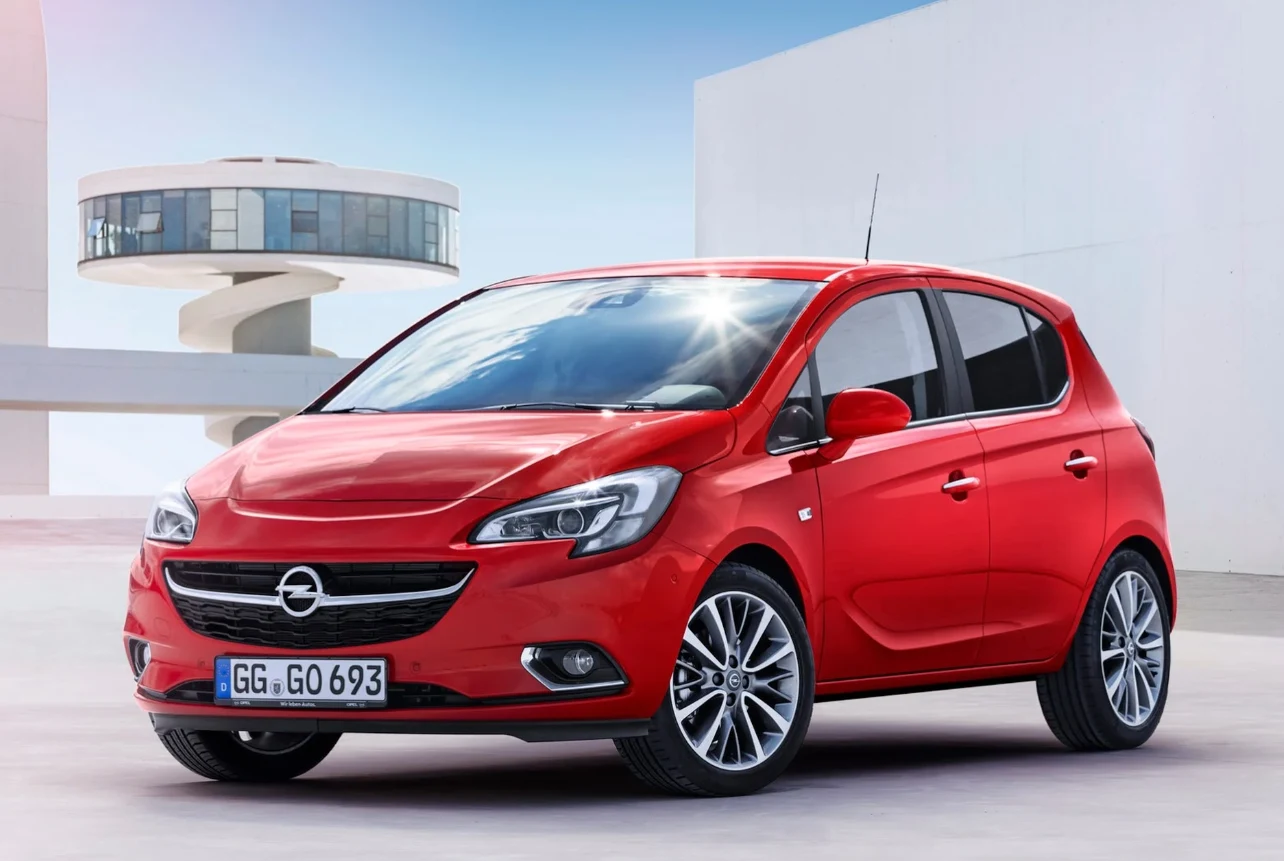 Litera E w nazwie nie oznacza wersji elektrycznej - Opel do oznaczania kolejnych generacji używa alfabetu, więc Corsa E to model produkowany w latach 2014-2019, następca generacji D i poprzednik generacji F. Czerwony Opel Corsa w wersji pięciodrzwiowej, ustawiony z przodu pod kątem, z charakterystycznym grillem i dynamicznymi reflektorami, na tle nowoczesnej, minimalistycznej architektury.