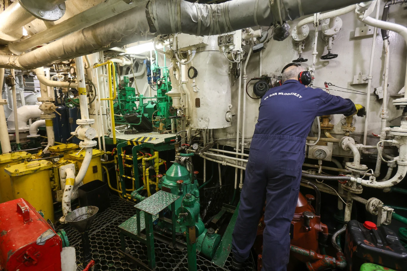 mechanik w kombinezonie i ochronnych słuchawkach pracuje przy obsłudze skomplikowanych urządzeń mechanicznych i rur w maszynowni statku, otoczony licznymi zaworami, przewodami i kolorowymi maszynami