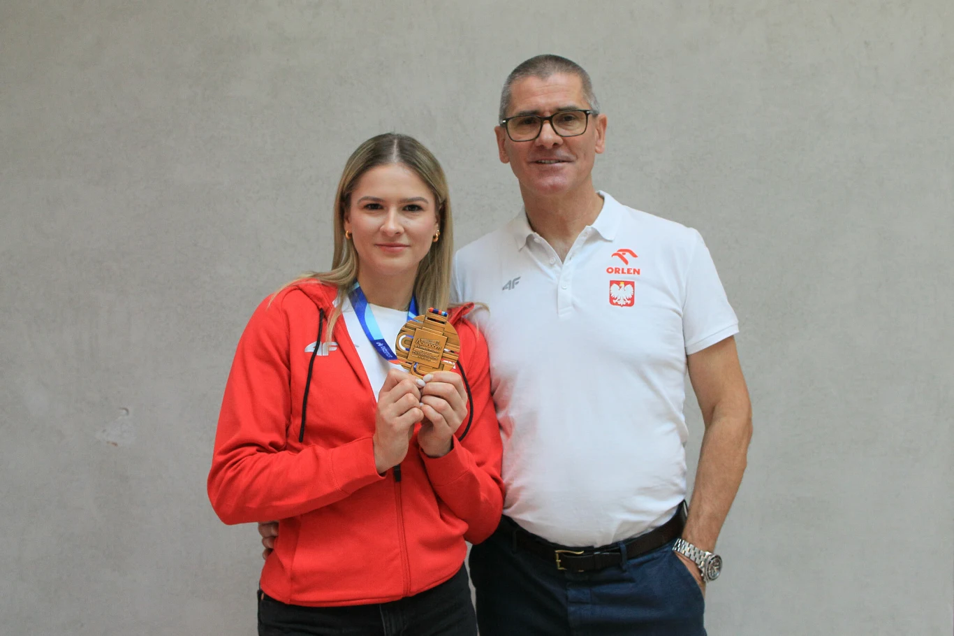 kobieta w czerwonej sportowej kurtce trzyma w dłoni złoty medal zawieszony na niebiesko-białej wstążce, obok niej stoi mężczyzna w białej koszulce z polskim godłem i napisem 'Polska'