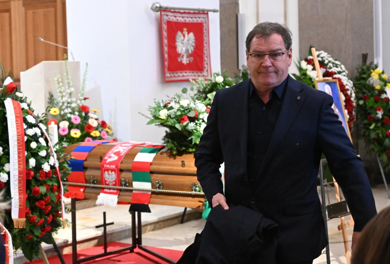 mężczyzna ubrany na czarno stoi obok trumny przykrytej flagami i szalikami, wokół liczne wieńce oraz biało-czerwona flaga z godłem Polski na ścianie