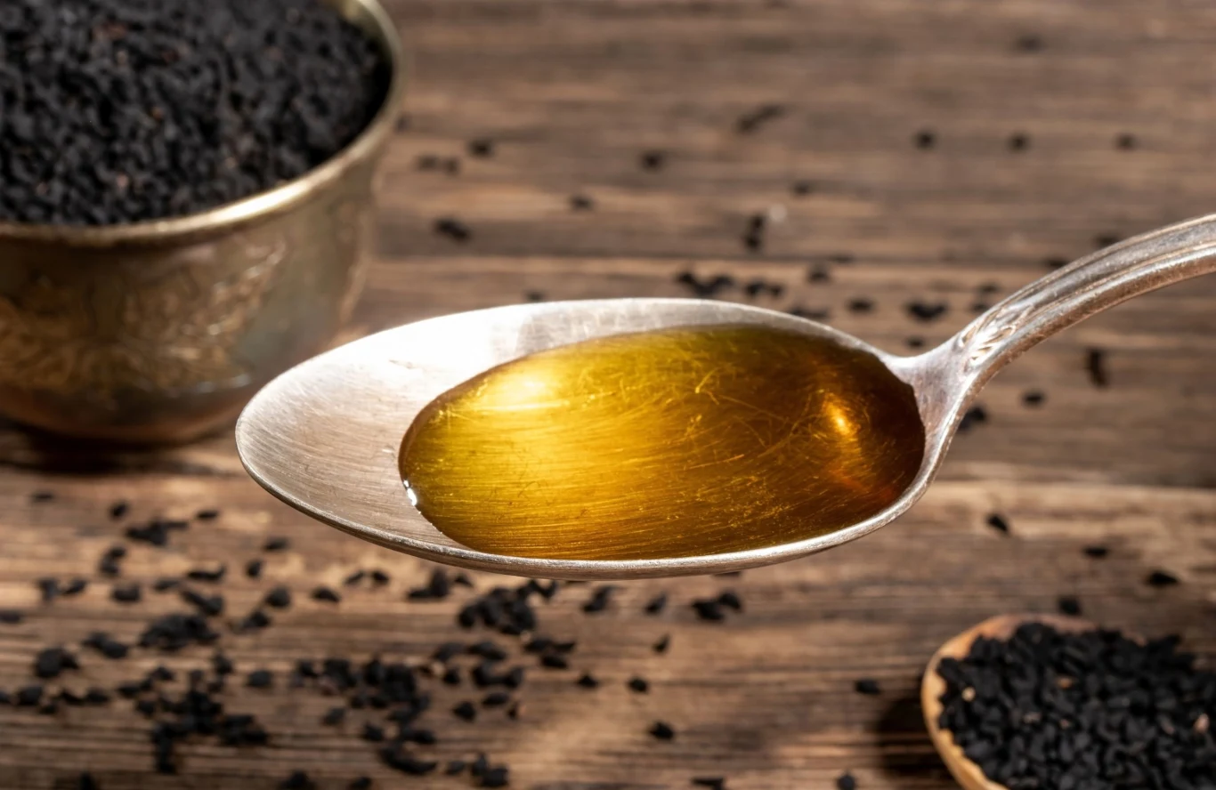 Olej z czarnuszki jest bogaty w zdrowe, nienasycone kwasy tłuszczowe Łyżka z olejem z czarnuszki na drewnianym stole, obok rozsypane i miska z nasionami czarnuszki.