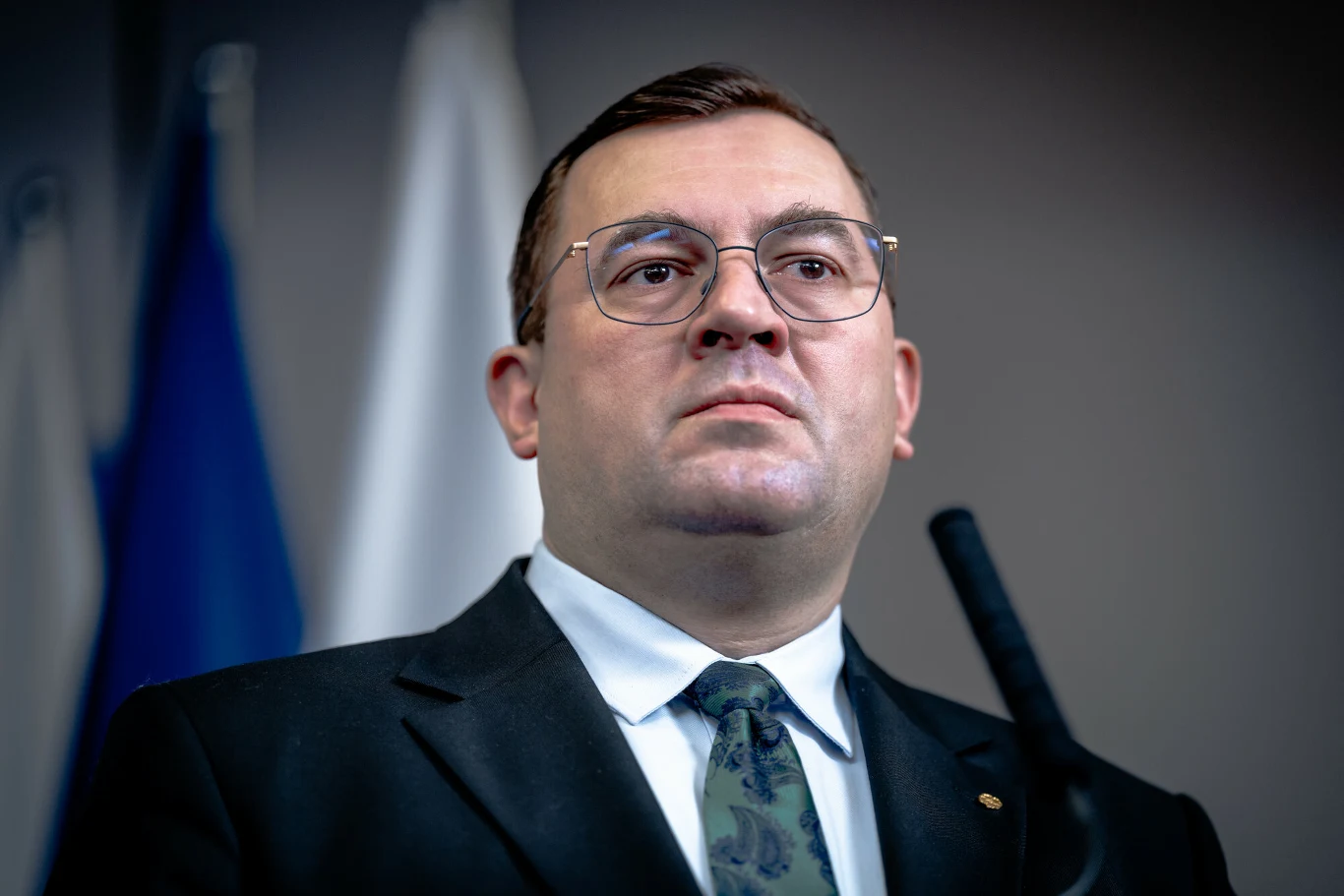 Stefan Krajewski. Stefan Krajewski w garniturze i okularach stoi przed mikrofonem, w tle rozmyte niebiesko-białe flagi.
