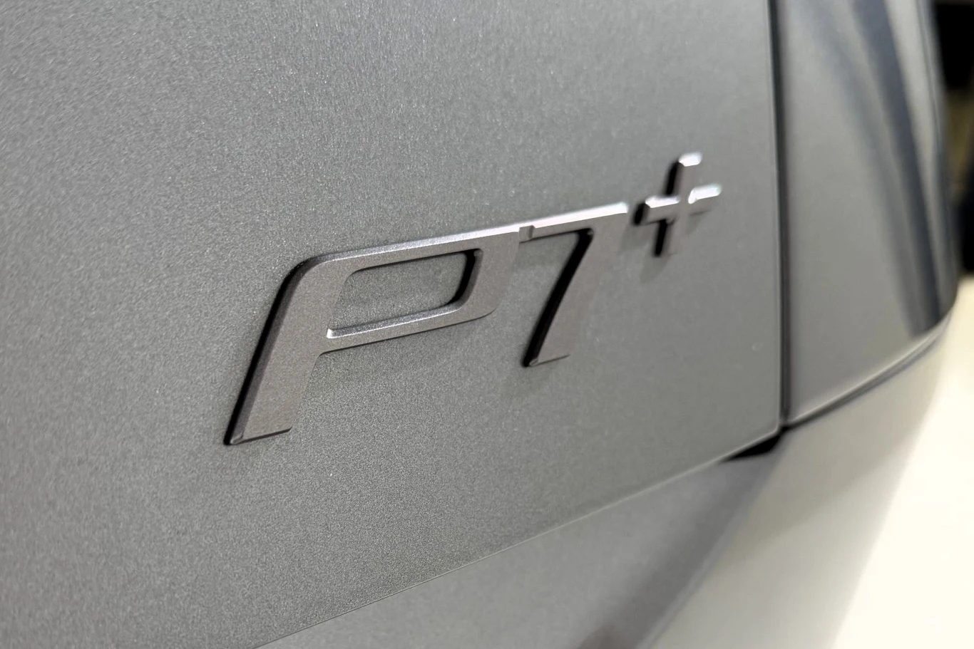 metaliczny emblemat z napisem „P7+” umieszczony na szarej, matowej karoserii samochodu