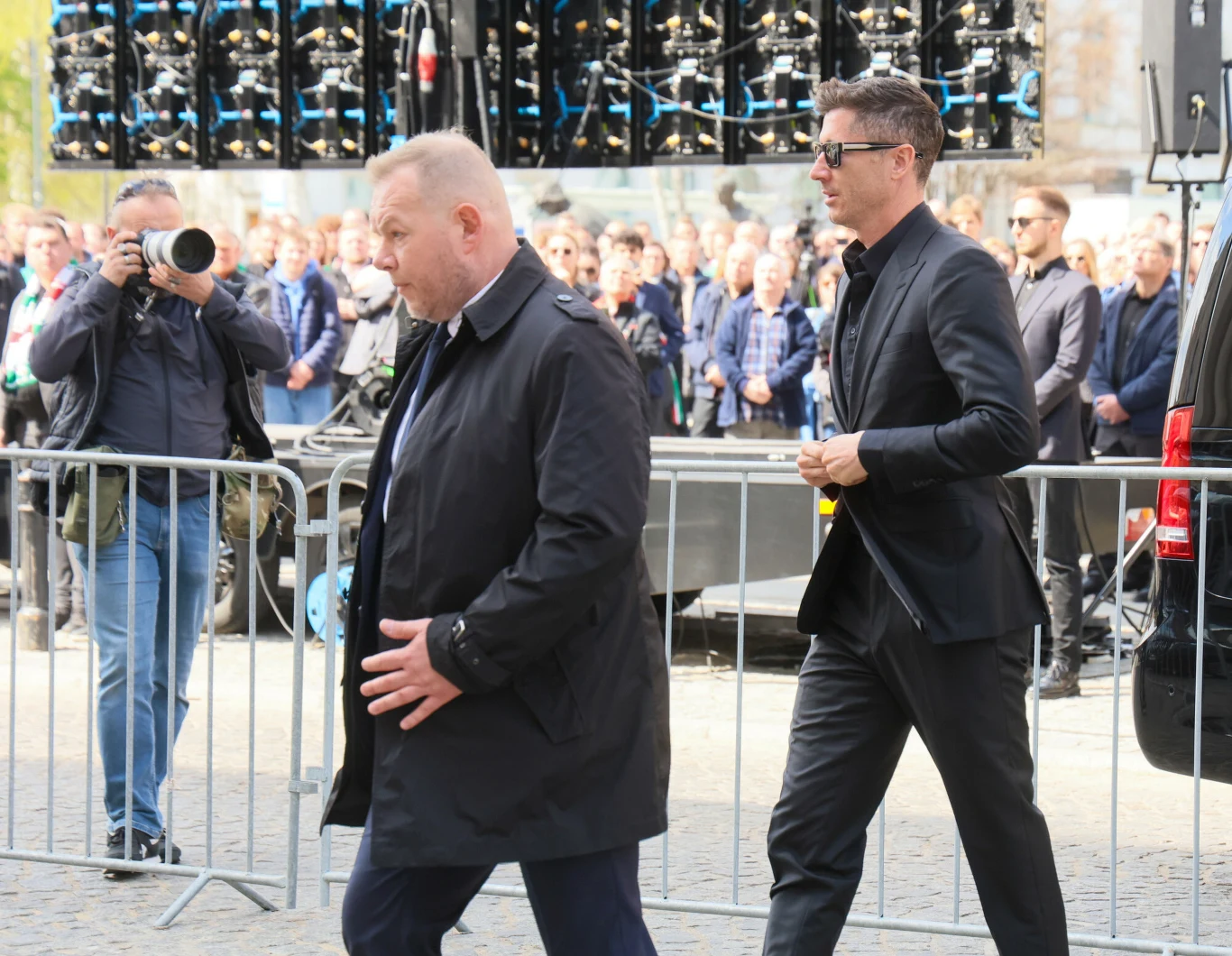 dwóch mężczyzn w ciemnych ubraniach idzie obok metalowych barierek, za nimi tłum ludzi oraz fotograf z aparatem, w tle widoczne reflektory sceniczne