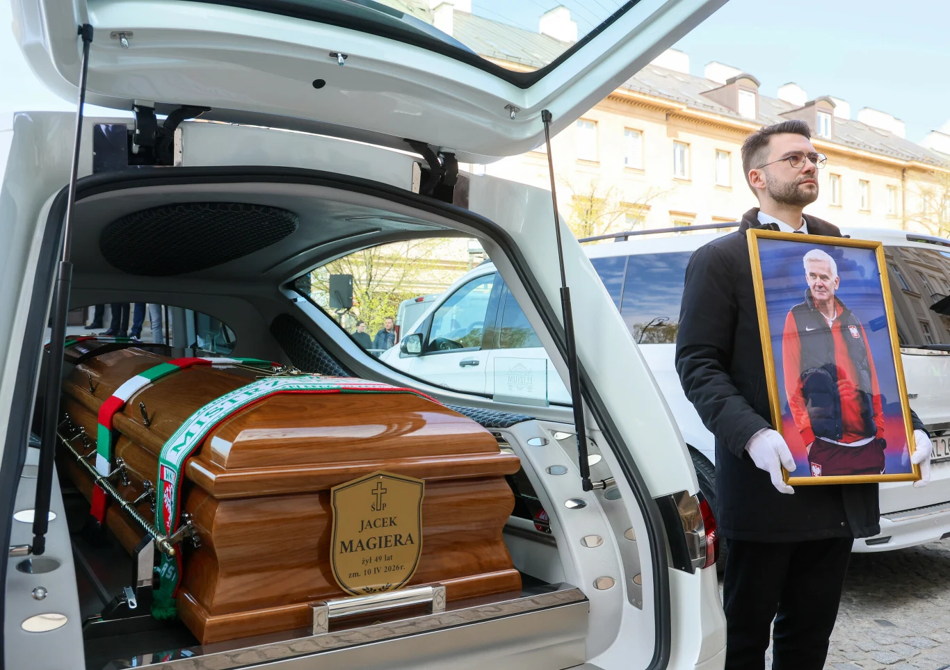 biała karawan pogrzebowy z otwartym bagażnikiem, w środku drewniana trumna z tabliczką z napisem 'Jacek Magiera', obok stoi mężczyzna w czarnym ubraniu i białych rękawiczkach trzymający duże zdjęcie starszego mężczyzny w kurtce