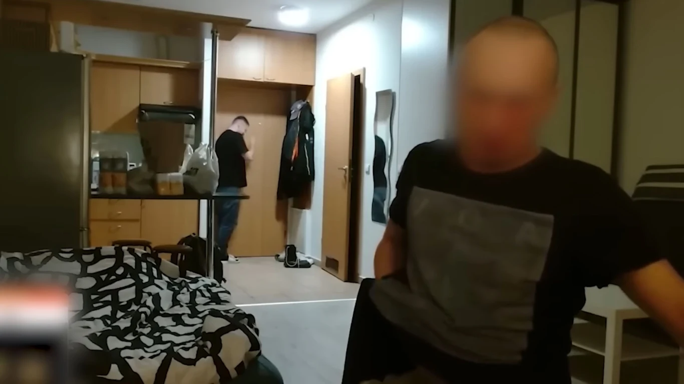 Patostreaming to transmitowanie w internecie ekstremalnych zachowań o charakterze wulgarnym, obscenicznym, szokującym lub brutalnym / źrodło: YouTube.com @Konopskyy Dwóch mężczyzn w mieszkaniu; jeden siedzi przodem do kamery z rozmazaną twarzą, drugi odwrócony tyłem stoi przy drzwiach wejściowych. W tle widoczna kuchnia i część łóżka z pościelą o czarno-białym wzorze.