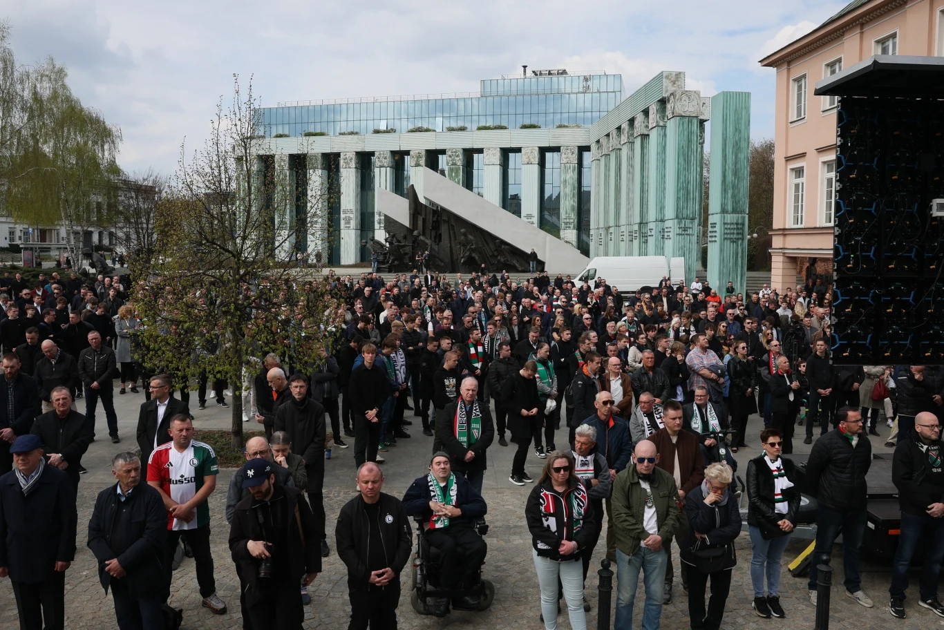 duży tłum ludzi zgromadzony przed pomnikiem Powstania Warszawskiego, wiele osób ubranych na ciemno, niektórzy noszą szaliki piłkarskie, w tle nowoczesny budynek z kolumnami oraz część starszej zabudowy