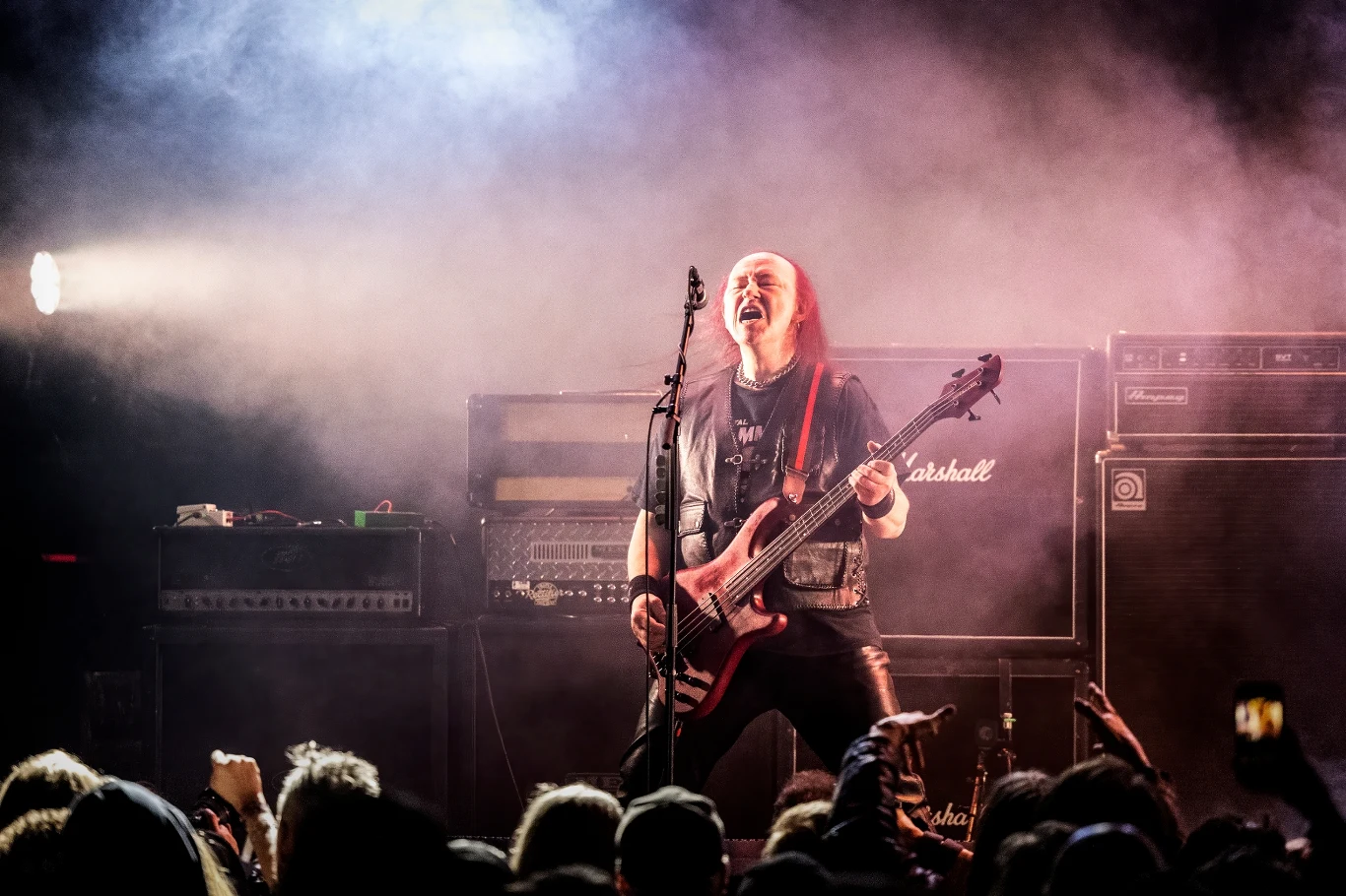 Cronos (Venom) w akcji Cronos, basista Venom, grający i śpiewający na oświetlonej scenie wśród mgły, widoczny tłum pod sceną