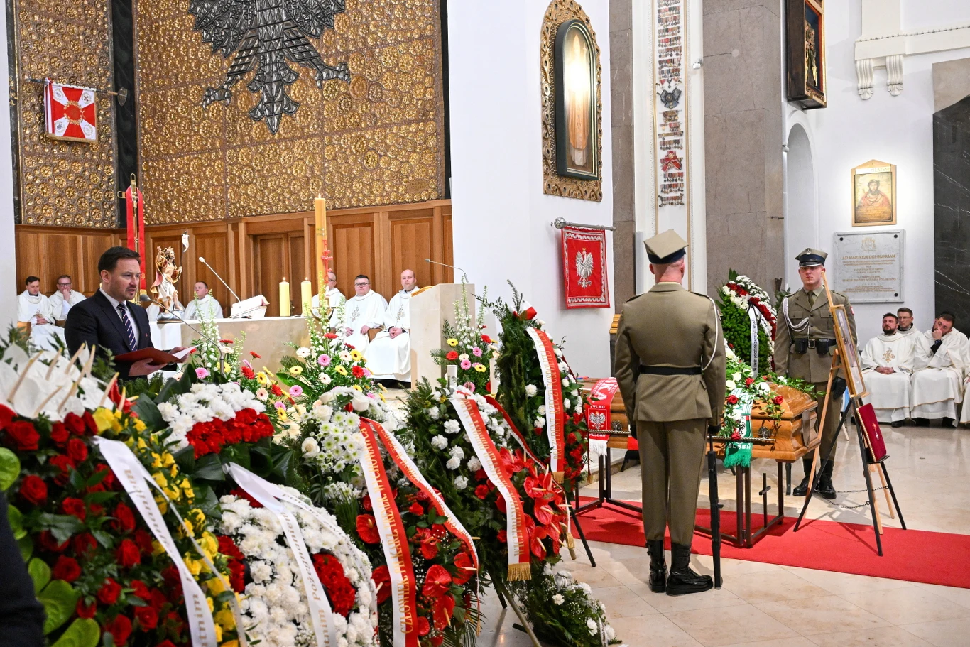 trumna przystrojona licznymi wieńcami kwiatów ustawiona w centralnej części kościoła, wartownicy w mundurach wojskowych stojący obok, a w tle grupa duchownych i mężczyzna przemawiający przy mównicy; ściany zdobią patriotyczne symbole i portrety