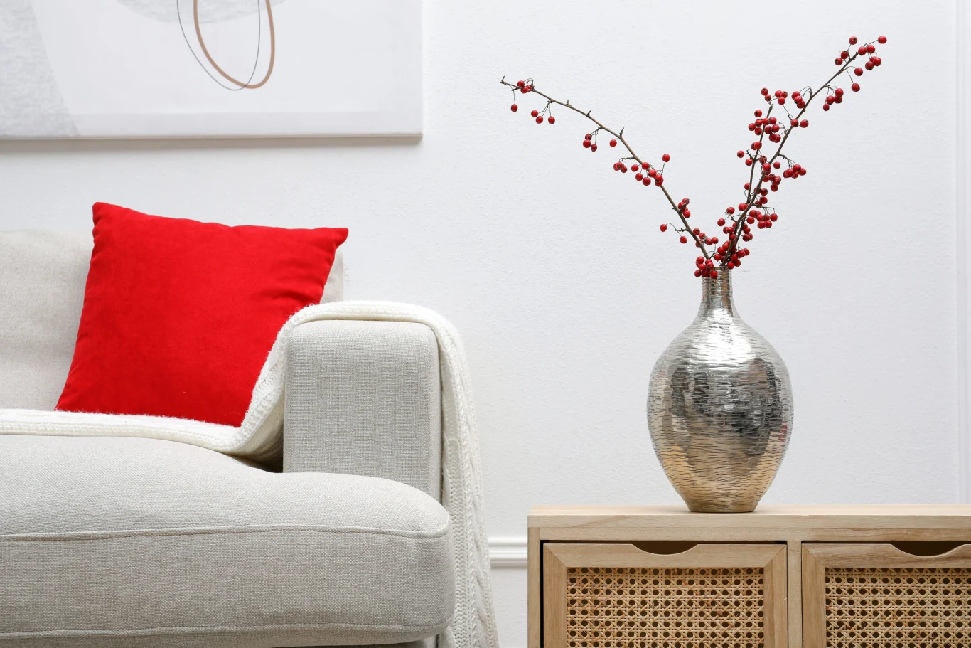Jasna sofa z wyrazistą czerwoną poduszką, obok drewniana szafka z metalicznym wazonem z gałązkami czerwonych jagód na białym tle ściany.