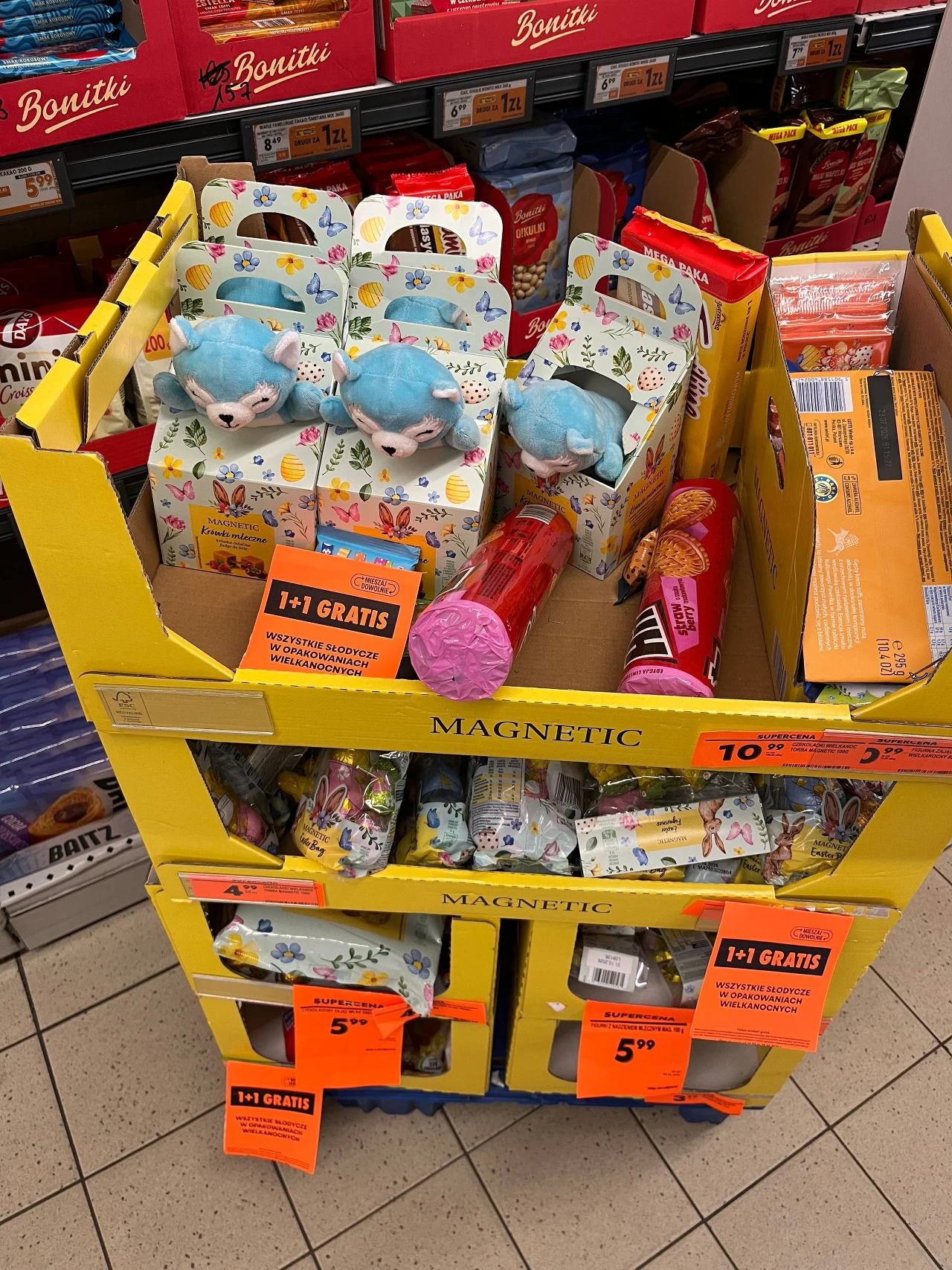 żółty regał sklepowy wypełniony kolorowymi zabawkami i słodyczami, z widoczną promocją 1+1 gratis na pomarańczowych etykietach, na górnej półce pluszowe maskotki z niebieskimi włosami oraz opakowania wielkanocne