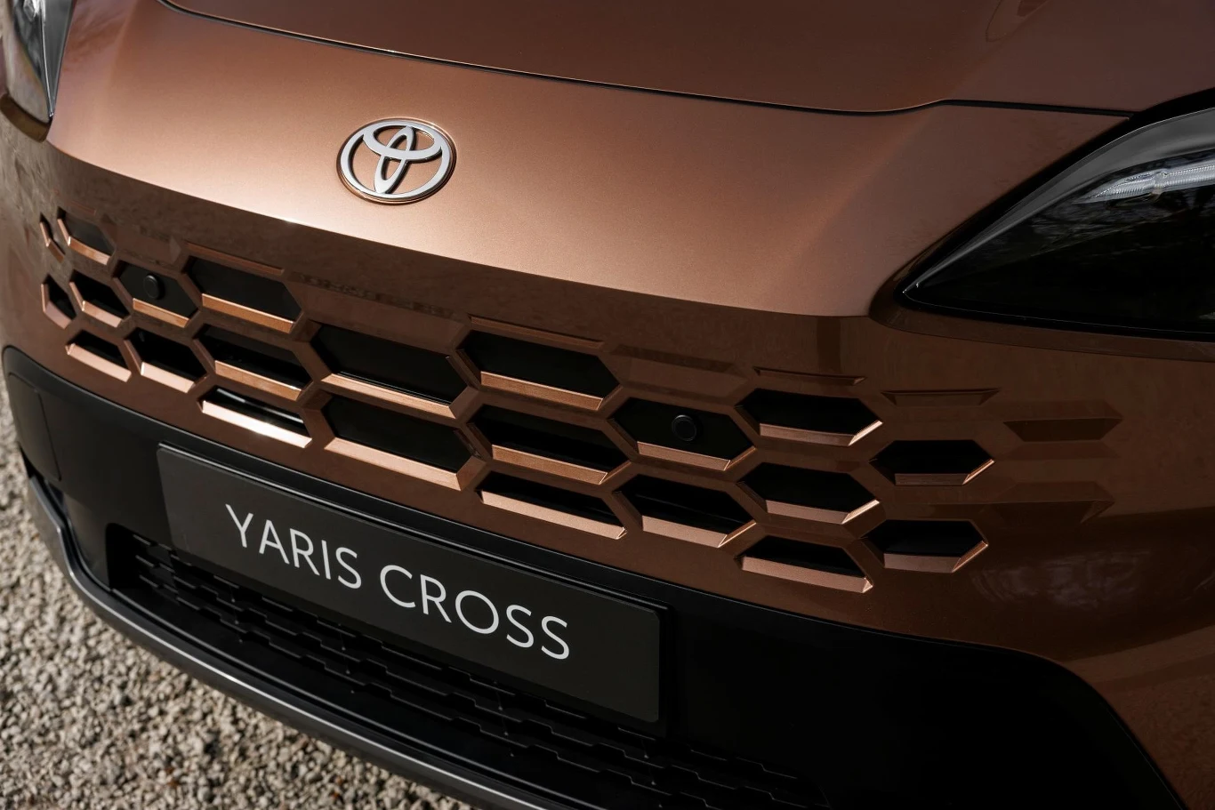 przedni grill Toyoty Yaris Cross w kolorze brązowym z wyraźnie zaznaczonym geometrycznym wzorem oraz logo producenta umieszczonym centralnie nad charakterystyczną tablicą z nazwą modelu