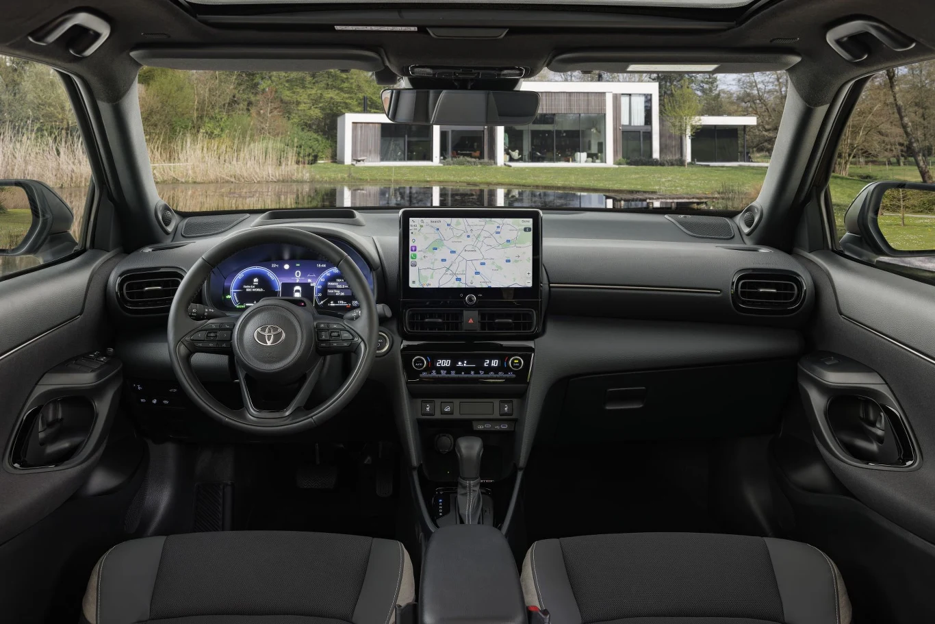 nowoczesna deska rozdzielcza samochodu Toyota z dużym centralnym ekranem dotykowym wyświetlającym mapę, cyfrowym zestawem wskaźników oraz panoramicznym widokiem na modernistyczny dom z dużymi przeszkleniami w tle