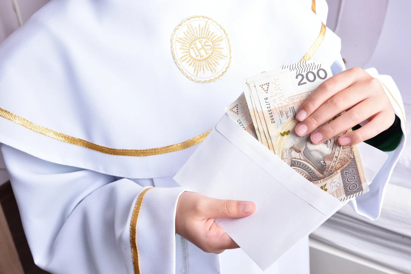 Kto ma prawo do zarządzania majątkiem z pierwszej komunii świętej? Osoba w białej albie z haftem wkłada banknoty 200 zł do białej koperty, temat: majątek z komunii