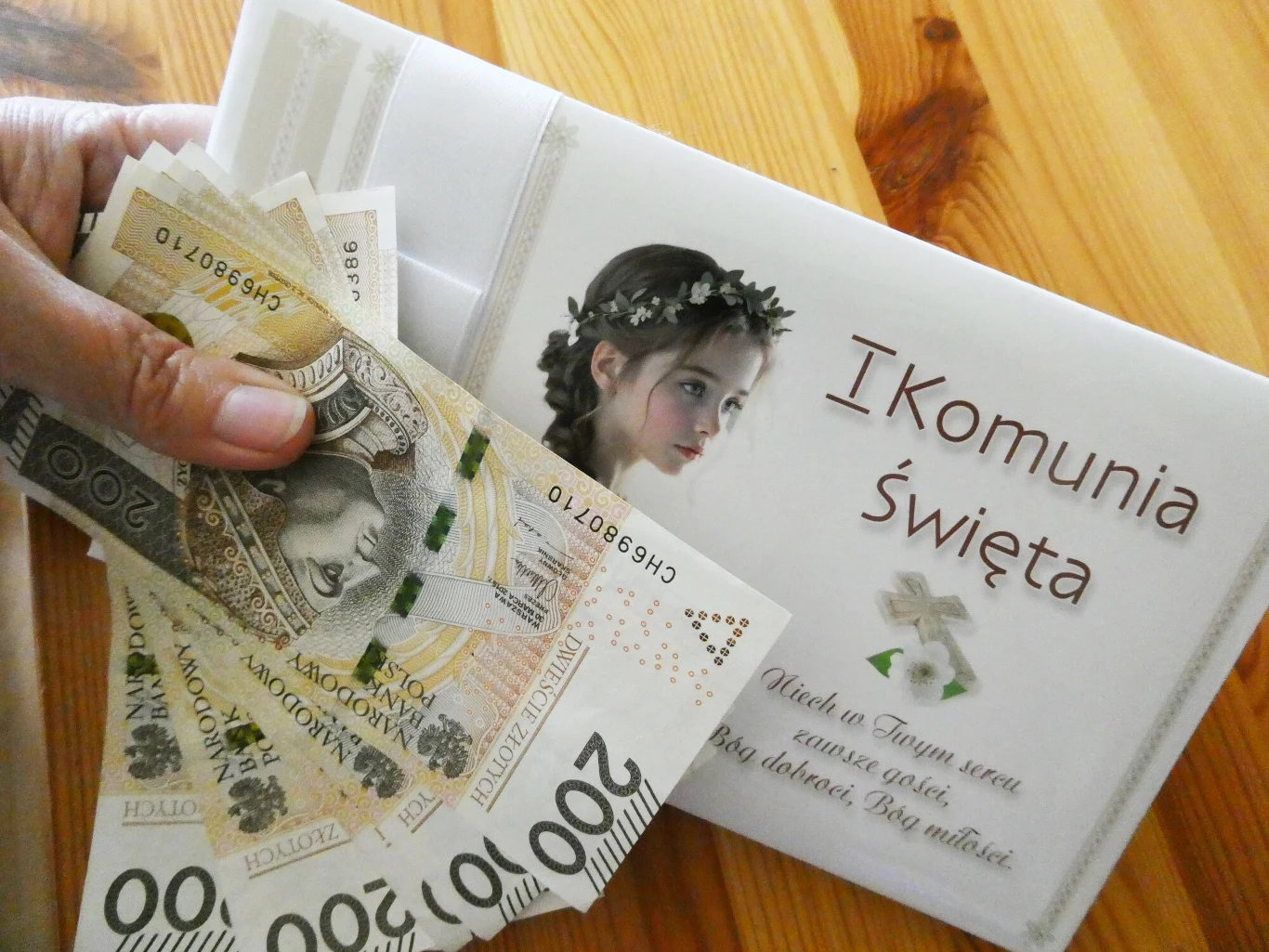 Rozsądnym rozwiązaniem w kontekście dysponowania i wydawania pieniędzy dziecka, jest podejmowanie wspólnych decyzji, na co zostaną one przeznaczone Koperta ozdobiona zdjęciem dziewczynki w wianku i napisem „I Komunia Święta” na stole, obok której ktoś trzyma wachlarz z banknotami o nominałach 100 i 200 złotych.