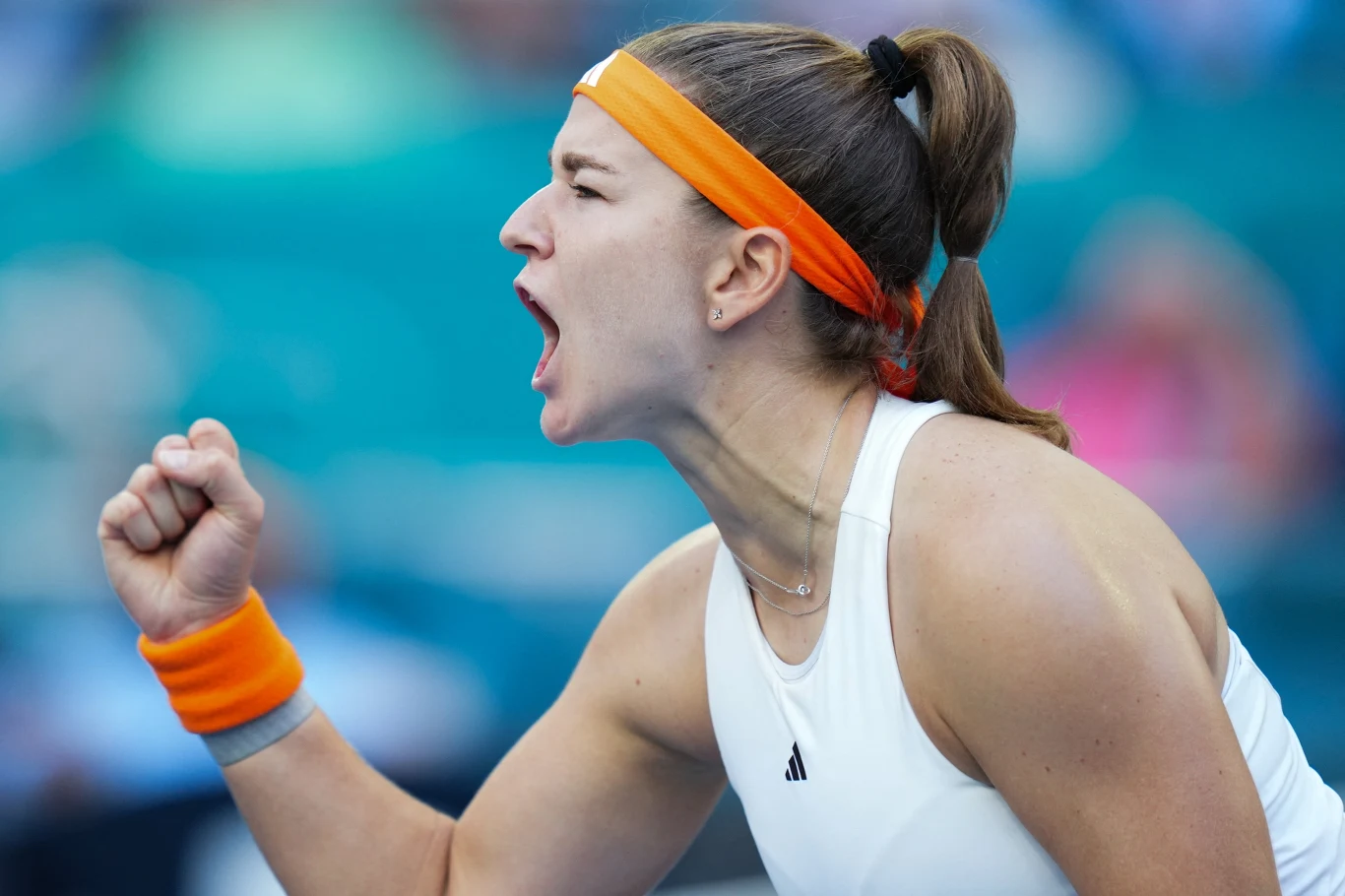 tenisistka w białym stroju i pomarańczowej opasce na głowie zaciska pięść i głośno wyraża emocje, ukazując determinację i sportową pasję