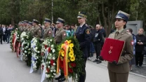szpaler żołnierzy i funkcjonariuszy w mundurach stojących z wieńcami z kwiatów oraz kobieta trzymająca odznaczenie w czerwonym etui, w tle zgromadzeni ludzie i drzewa