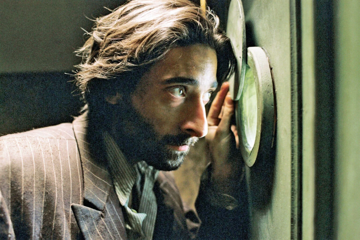 Adrien Brody w scenie z "Pianisty" Adrien Brody w "Pianiście" zagląda przez okrągłe okienko w drzwiach, w marynarce w prążki, oświetlony światłem.