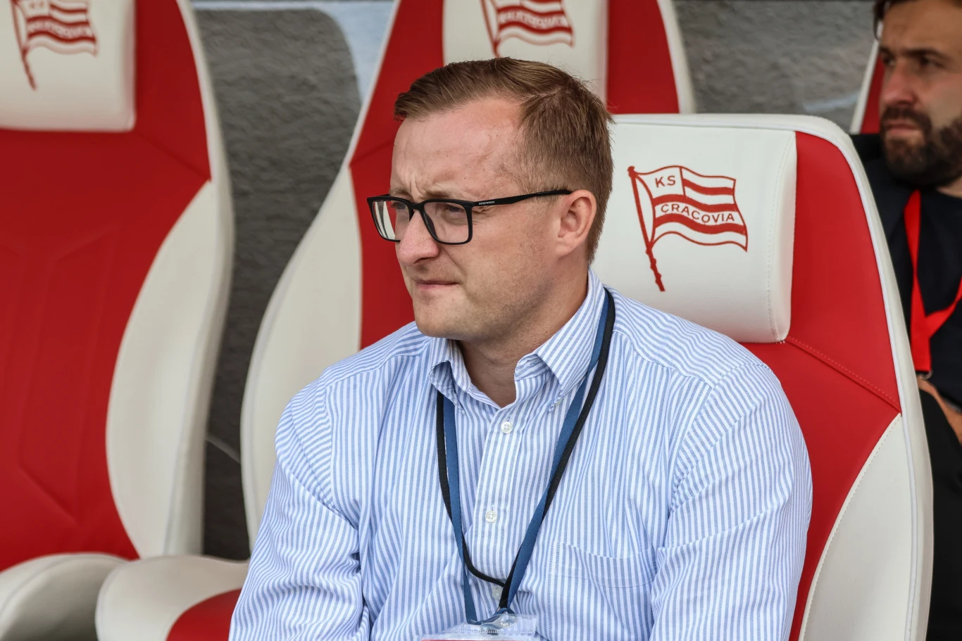Mężczyzna w okularach i koszuli w niebieskie paski siedzi na fotelu z logo KS Cracovia, obok niego inny mężczyzna z brodą, również na tym samym tle klubowym.