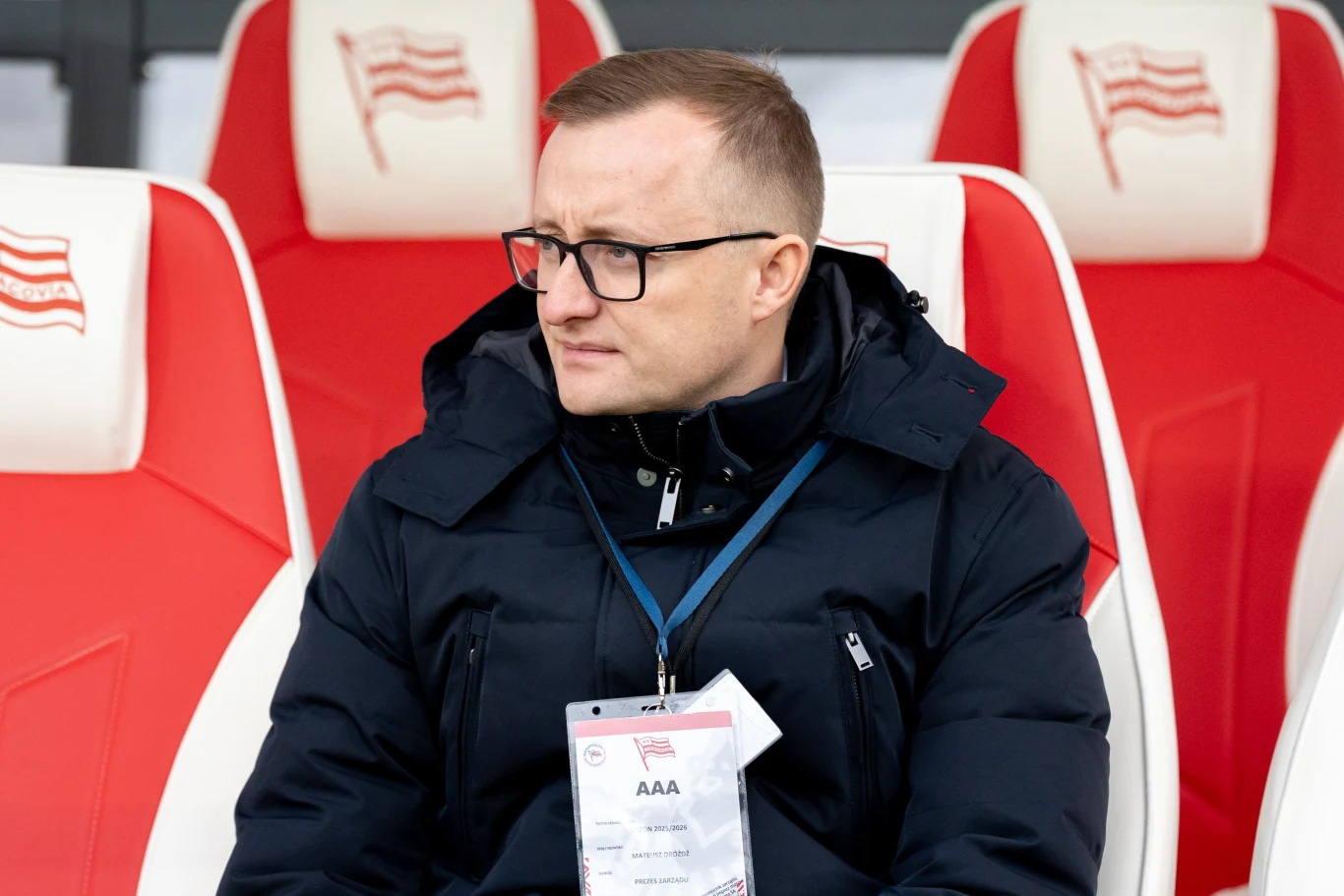 mężczyzna w okularach i ciemnej kurtce siedzi na stadionowej ławce z czerwono-białymi fotelami ozdobionymi flagami, na szyi zawieszony identyfikator AAA