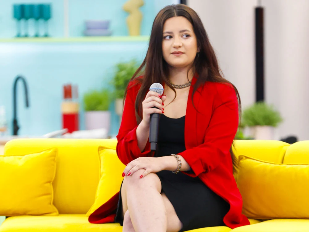 Oliwia Yanez wystąpi w programie "Must Be The Music" Oliwia Yanez w czerwonej marynarce i czarnej sukience siedzi na żółtej sofie z mikrofonem; jasne wnętrze, rośliny.