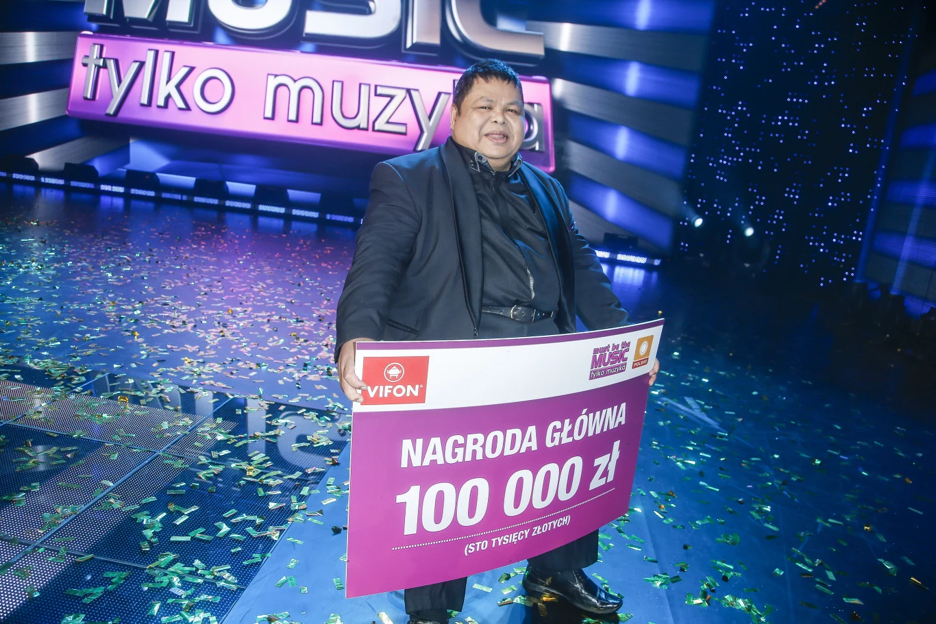 Conrado Yanez w 2015 r. wygrał w programie "Must Be The Music" uśmiechnięty mężczyzna ubrany w ciemny garnitur trzyma dużą planszę z napisem 'nagroda główna 100 000 zł', wokół leży konfetti, w tle widoczne neony i scenografia telewizyjna z logo programu 'tylko muzyka'