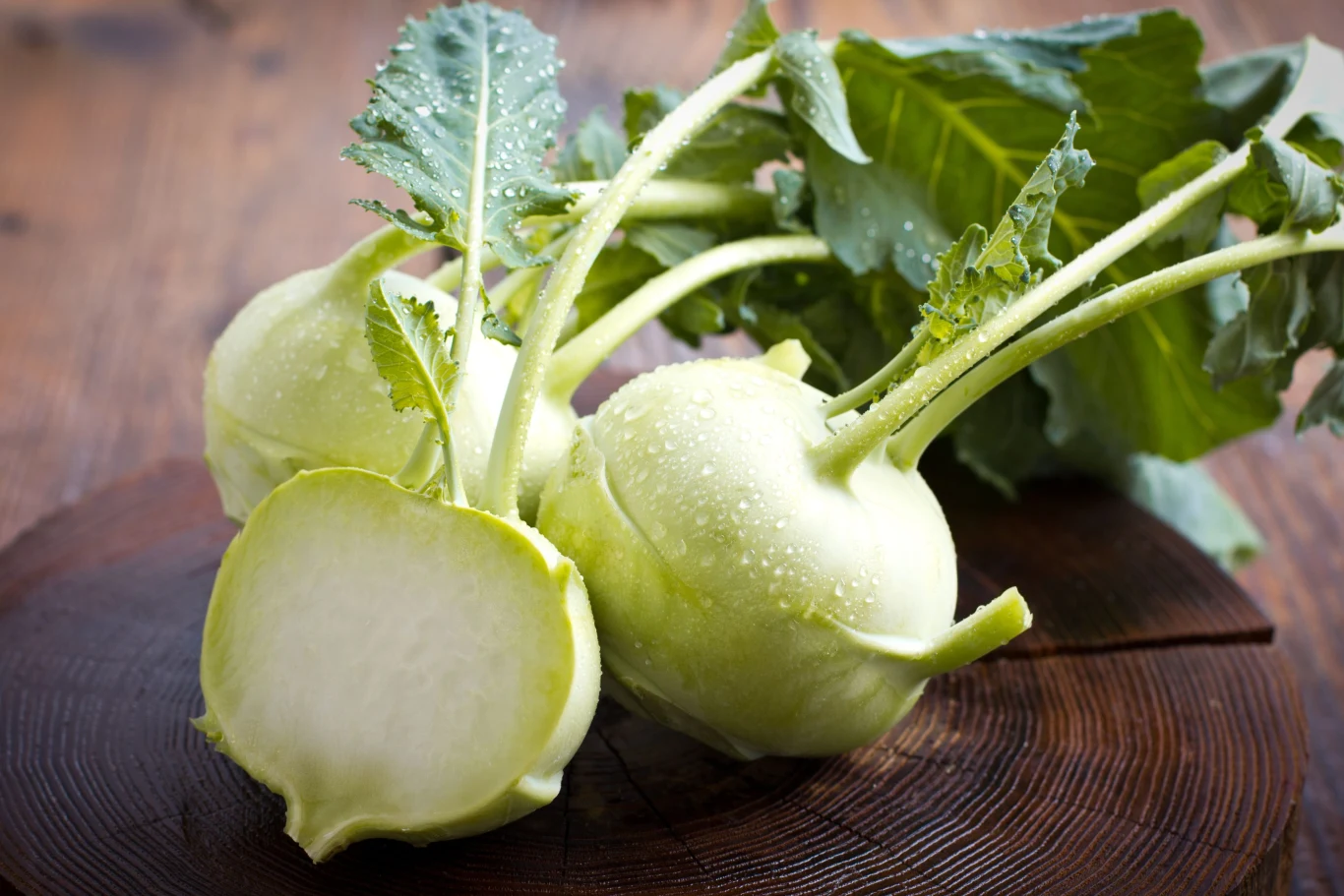 Dwie świeże kalarepy z zielonymi liśćmi, jedna z nich przekrojona na pół, leżą na drewnianej desce na tle rozmytego brązowego stołu.