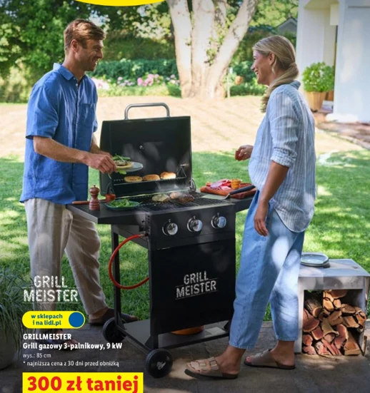 dwoje ludzi grilluje na czarnym grillu gazowym Grill Meister, na którym leżą warzywa i mięso, w tle zielony ogród, po prawej stronie stos drewna opałowego, na dole z przodu żółto-czerwony napis informujący o promocji 300 zł taniej