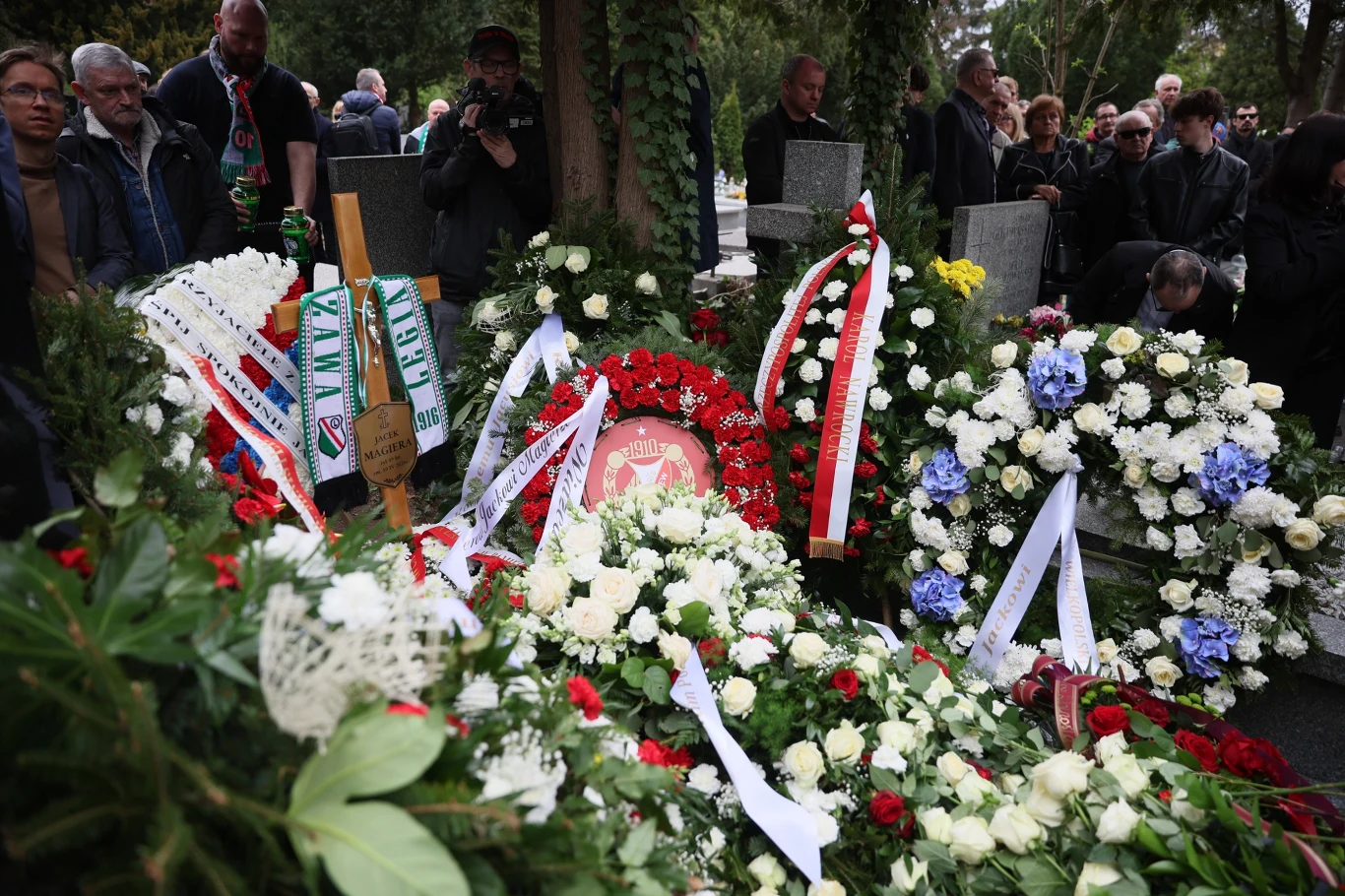 grób udekorowany licznymi wieńcami i kwiatami, dominują biało-czerwone kompozycje kwiatowe oraz szarfy narodowe, w otoczeniu zgromadzeni ludzie wyrażający żałobę