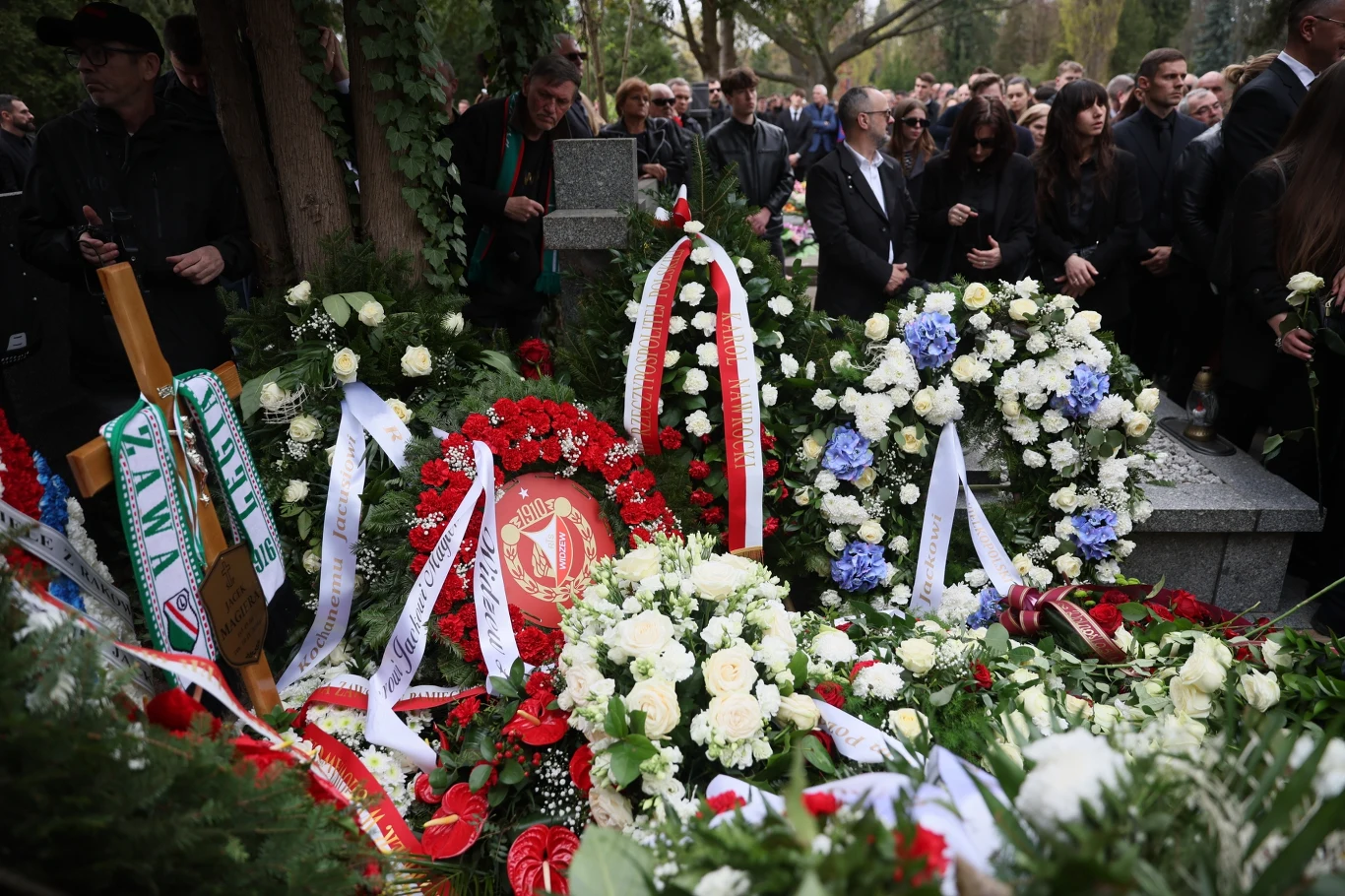 Grób Jacka Magiery Grób udekorowany licznymi wieńcami i kwiatami, w otoczeniu żałobników ubranych na czarno, skupionych wokół miejsca pochówku w cieniu drzew.