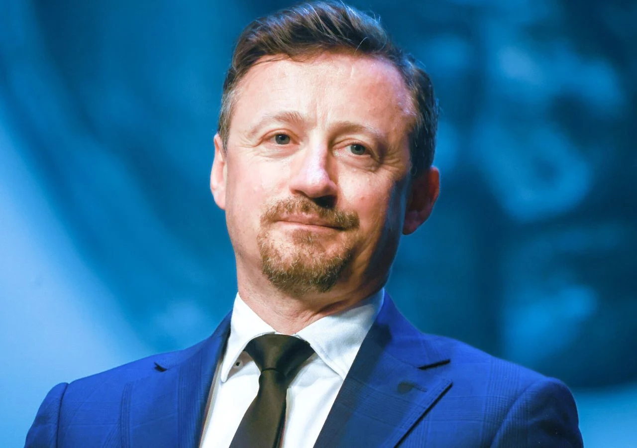 Adam Małysz Mężczyzna w średnim wieku z krótkimi brązowymi włosami i brodą, ubrany w niebieski garnitur, białą koszulę i czarny krawat, stoi na tle w odcieniach niebieskiego.