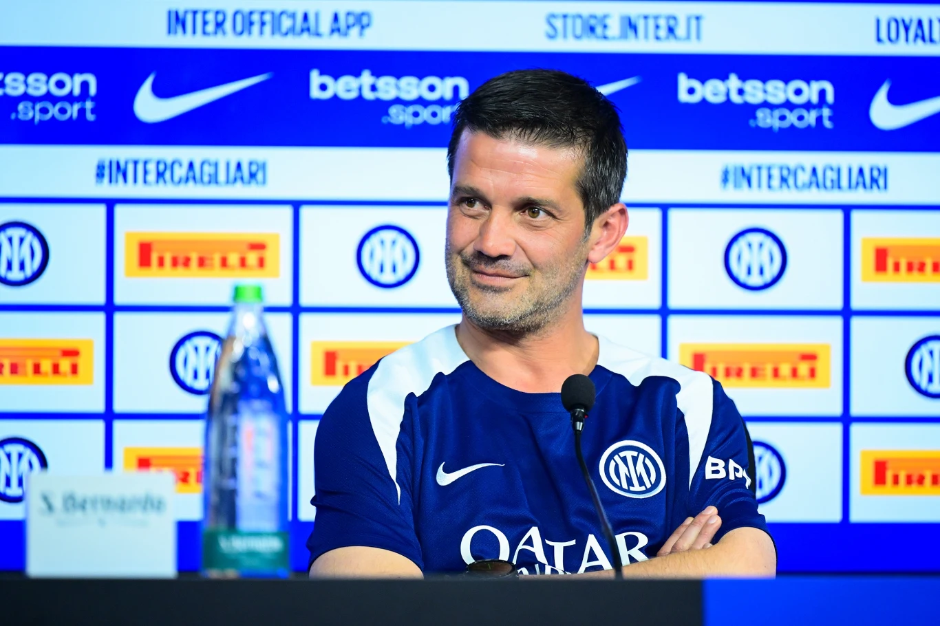 Cristian Chivu Trener piłkarski siedzi przy stole konferencyjnym z mikrofonem, ubrany w koszulkę Interu Mediolan, w tle widoczne są logotypy sponsorów i klubu na niebiesko-białej ściance medialnej, na stole stoi butelka wody i tabliczka z nazwiskiem.