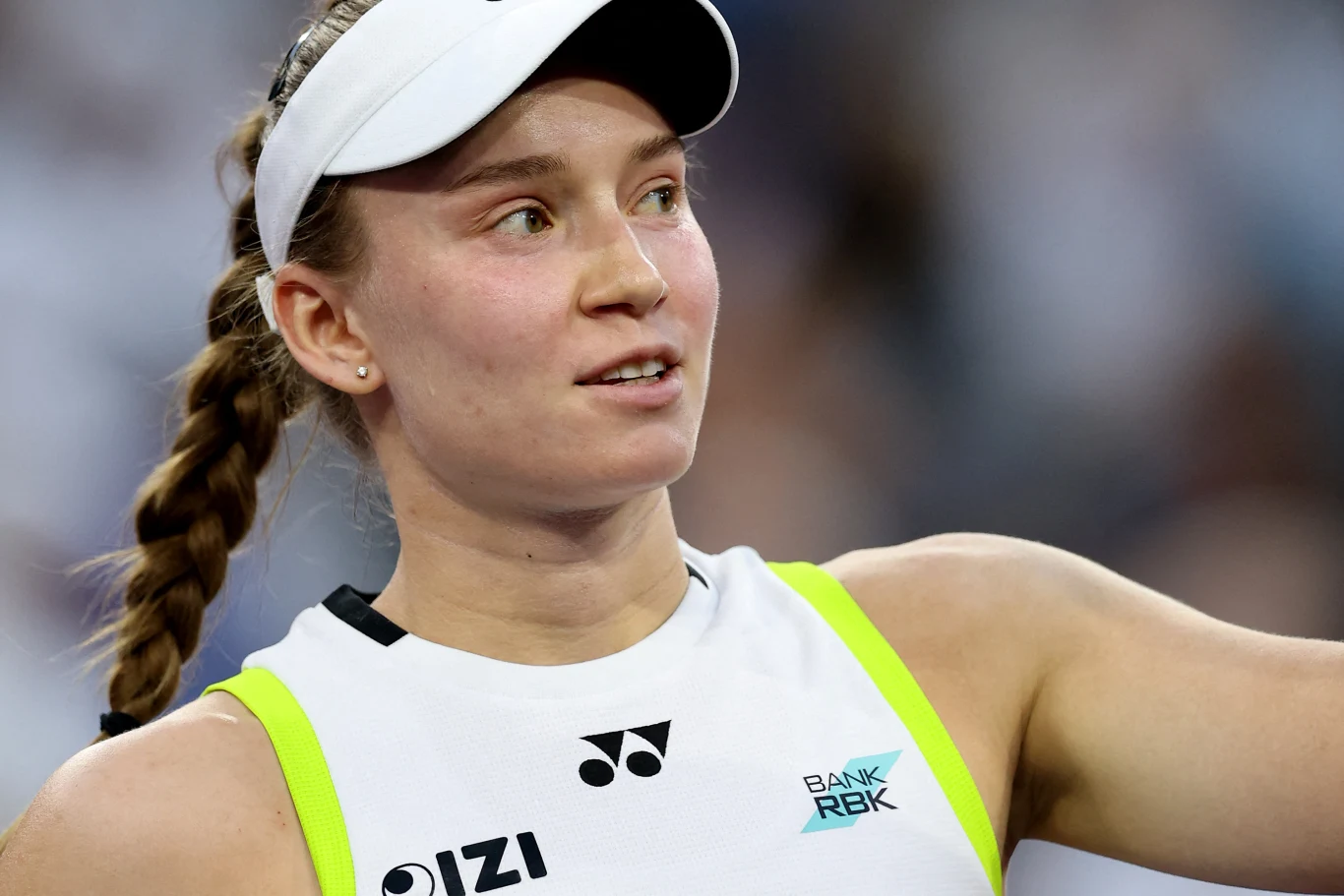 Młoda kobieta w sportowej czapce z daszkiem i białym topie z żółtymi akcentami, z widocznymi logotypami sponsorów, spogląda w bok z lekkim uśmiechem na twarzy, w tle rozmyta publiczność.