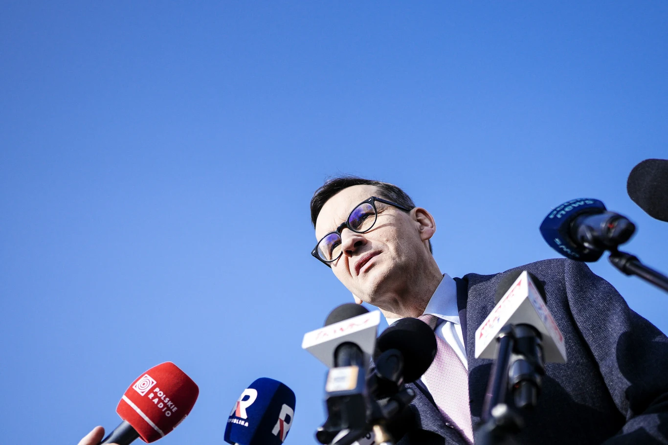 mężczyzna w okularach udziela wywiadu przed kilkoma mikrofonami różnych stacji telewizyjnych na tle jasnego nieba