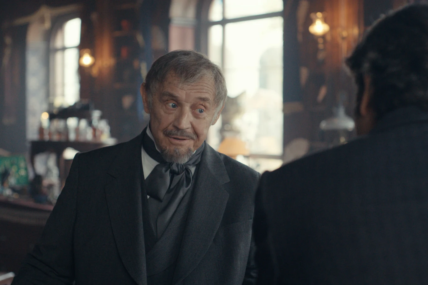 Kadr z filmu "Lalka" (2026) starszy mężczyzna w eleganckim, ciemnym garniturze z muchą prowadzi rozmowę z inną osobą w przytulnym, słabo oświetlonym wnętrzu, z widocznym w tle regałem pełnym butelek oraz dużym oknem