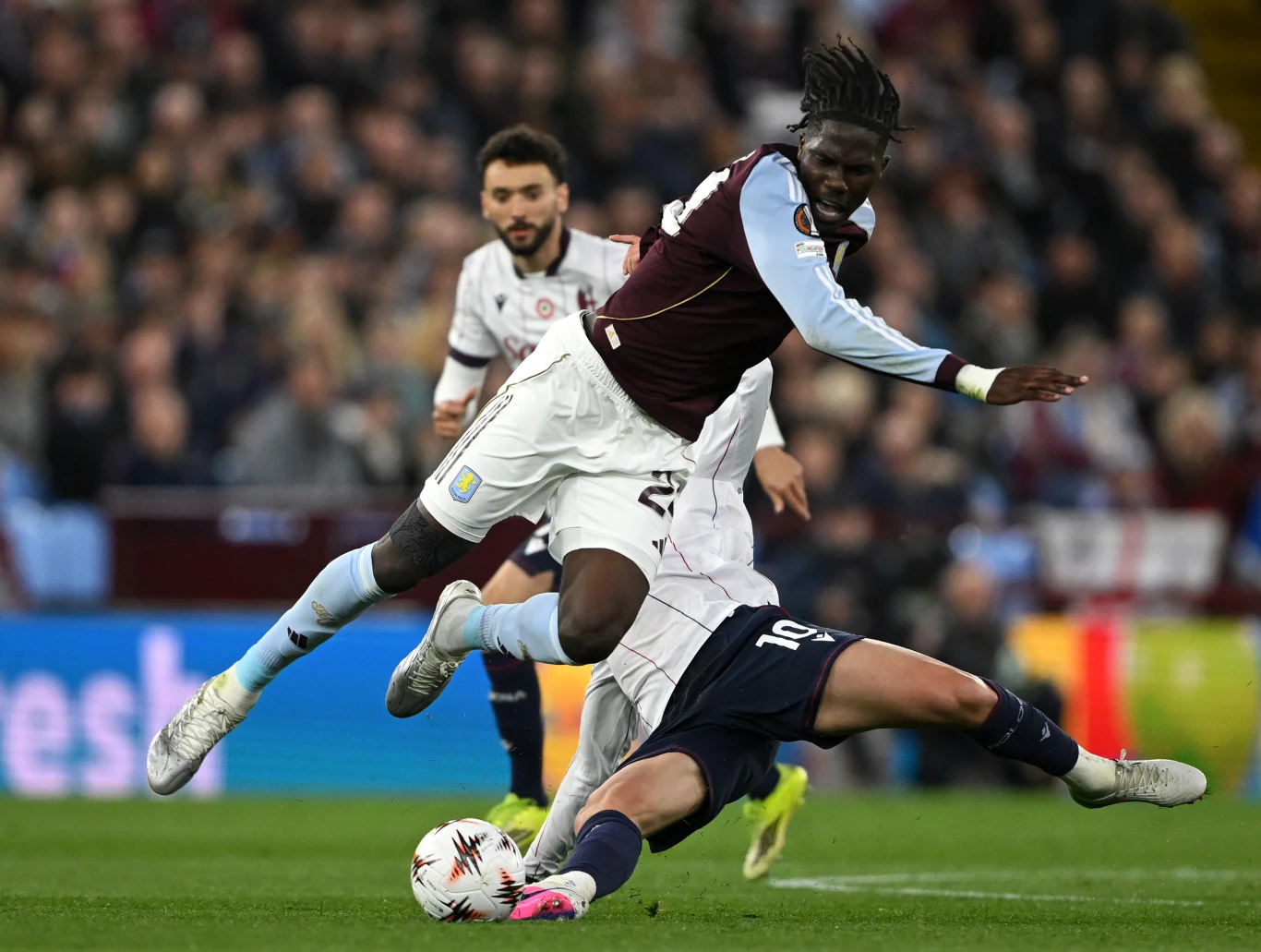 Kadr z meczu Aston Villa - Bologna piłkarz w bordowo-niebieskim stroju przeskakuje nad przeciwnikiem leżącym na murawie, podczas dynamicznej akcji meczowej na tle zatłoczonego stadionu