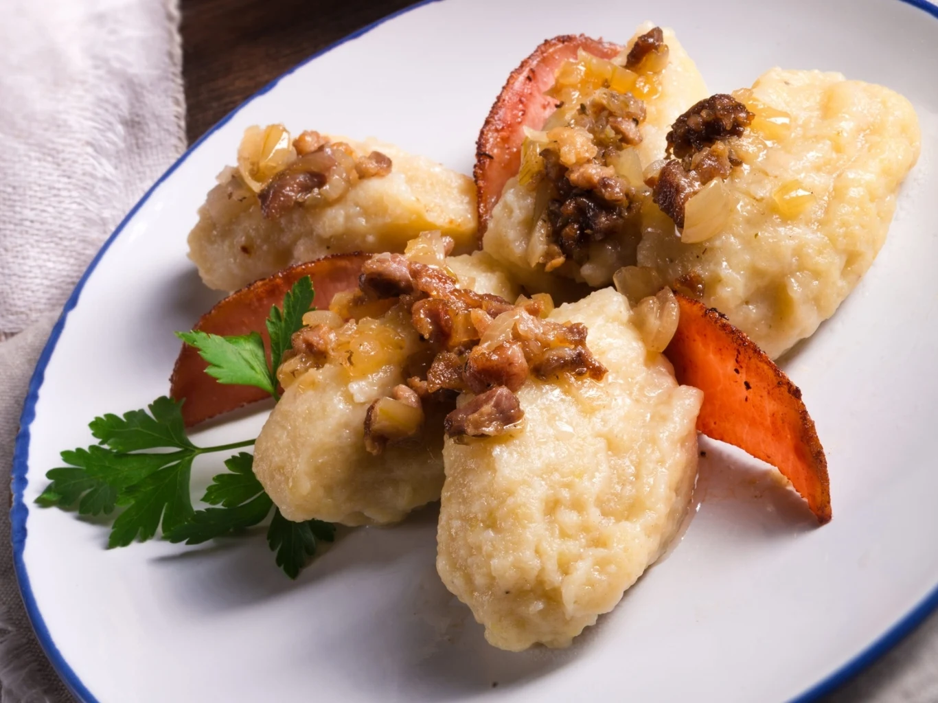 Kluski leniwe posypane podsmażoną cebulką z chrupiącymi skwarkami, udekorowane świeżą natką pietruszki oraz plasterkami boczku na białym talerzu.