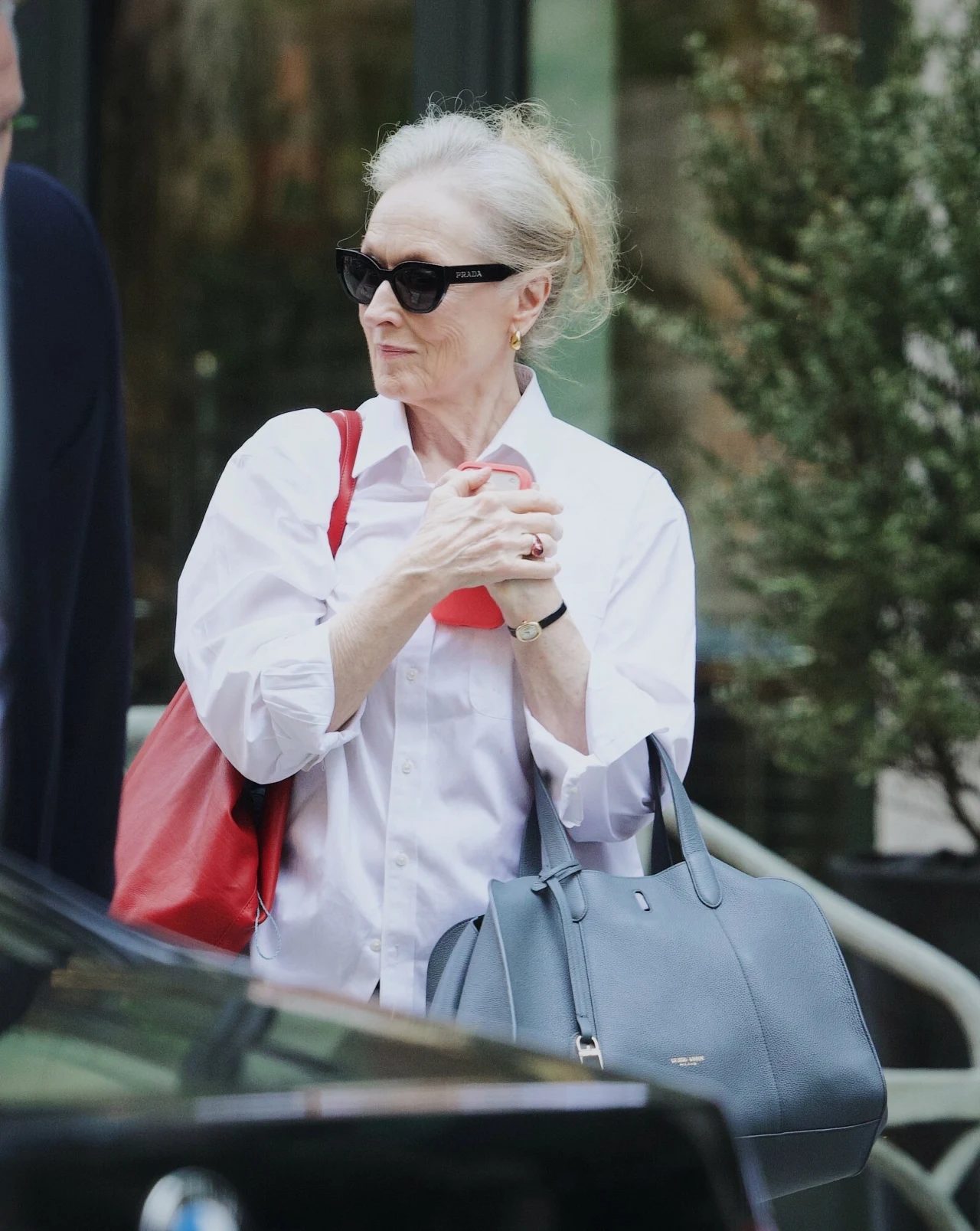 Meryl Streep zachwyca porcelanową cerą. Unika nadmiaru kosmetyków i pamięta o jednej zasadzie kobieta w białej koszuli i okularach przeciwsłonecznych trzyma dwie torebki, jedną czerwoną na ramieniu i drugą szarą w dłoni, stoi na tle zieleni i przeszklonego budynku
