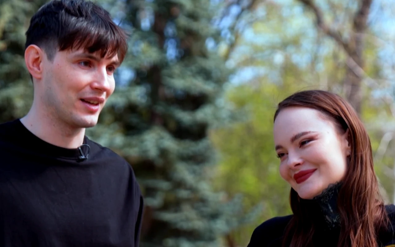 Dwie uśmiechnięte osoby rozmawiają ze sobą na tle zielonych drzew w słoneczny dzień, obie ubrane na czarno.