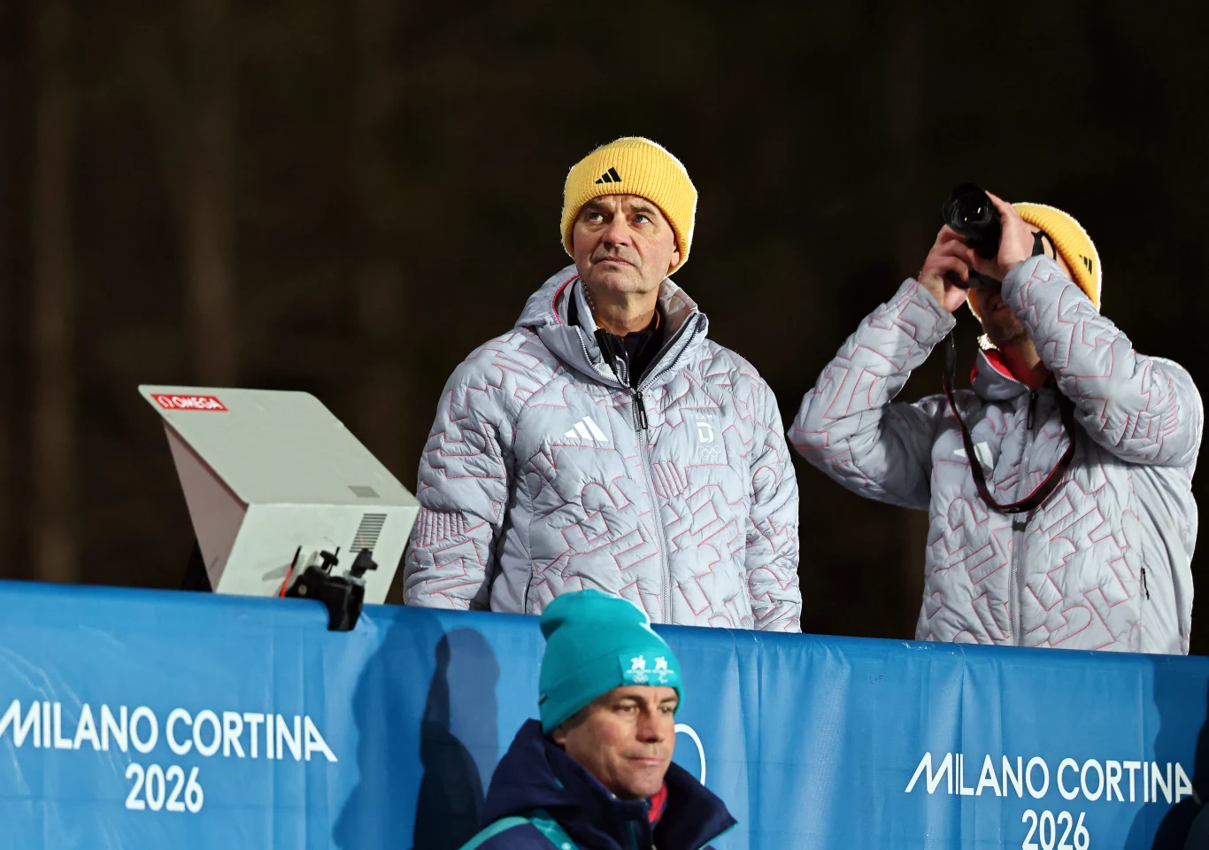 Dwóch mężczyzn w żółtych czapkach i srebrnych kurtkach stoi za niebieską barierką z napisem 'Milano Cortina 2026'; jeden z nich trzyma lornetkę przy oczach, a obok znajduje się kamera lub sprzęt elektroniczny, z przodu widać osobę w niebieskiej czapce.