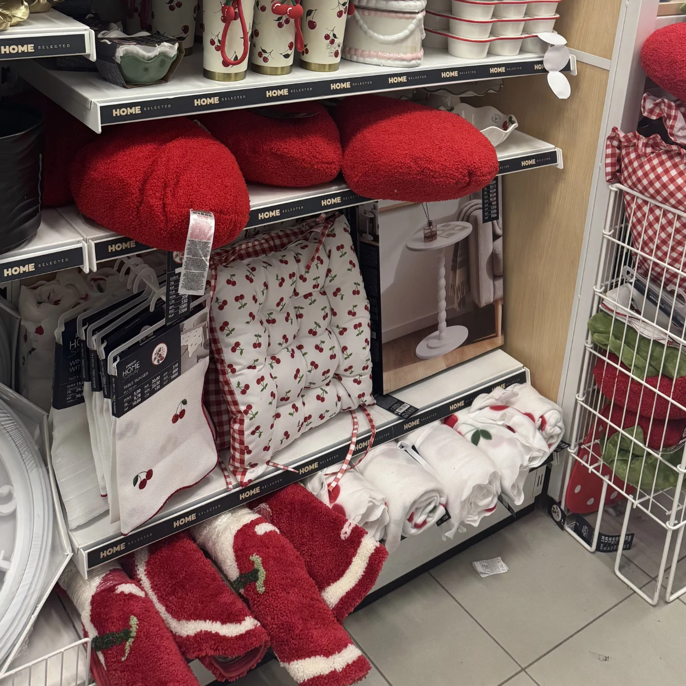 kolekcja tekstyliów domowych w odcieniach czerwieni i bieli oraz z motywem wiśni, w tym poduszki, ręczniki, ściereczki kuchenne i serwetki starannie poukładane na półkach w sklepie