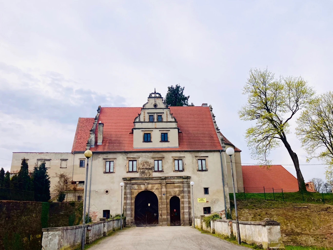 Brama do zamku na co dzień jest zamknięta, ale 2 maja otworzy się dla wszystkich zainteresowanych zwiedzaniem ruin zabytkowy budynek z czerwonym dachem, ozdobnym portalem wejściowym oraz symetrycznymi oknami, otoczony niskim murem i zielenią; po prawej stronie dwie smukłe drzewa bez liści