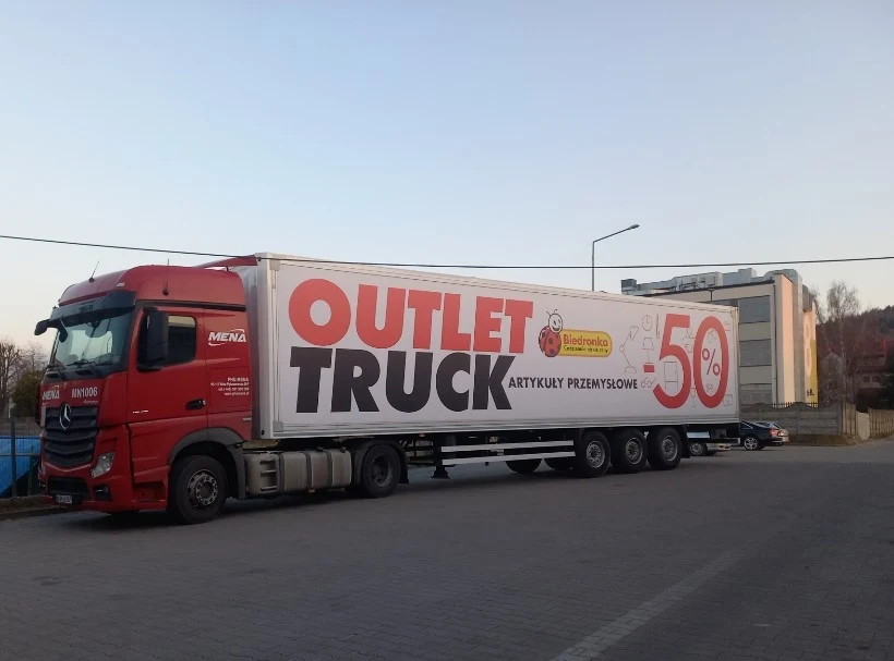 czerwony ciągnik siodłowy z naczepą, na której widnieje duży baner reklamowy Biedronki z napisem „OUTLET TRUCK ARTYKUŁY PRZEMYSŁOWE 50%”, zaparkowany na utwardzonym placu przy budynku