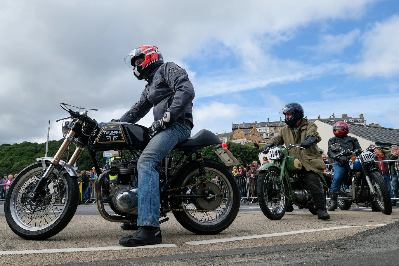 Trzech motocyklistów w kaskach stoi na swoich klasycznych motocyklach na poboczu ulicy, gotowych do startu; wokół nich zgromadził się tłum widzów, a w tle widoczne są budynki i fragment zielonego wzgórza.