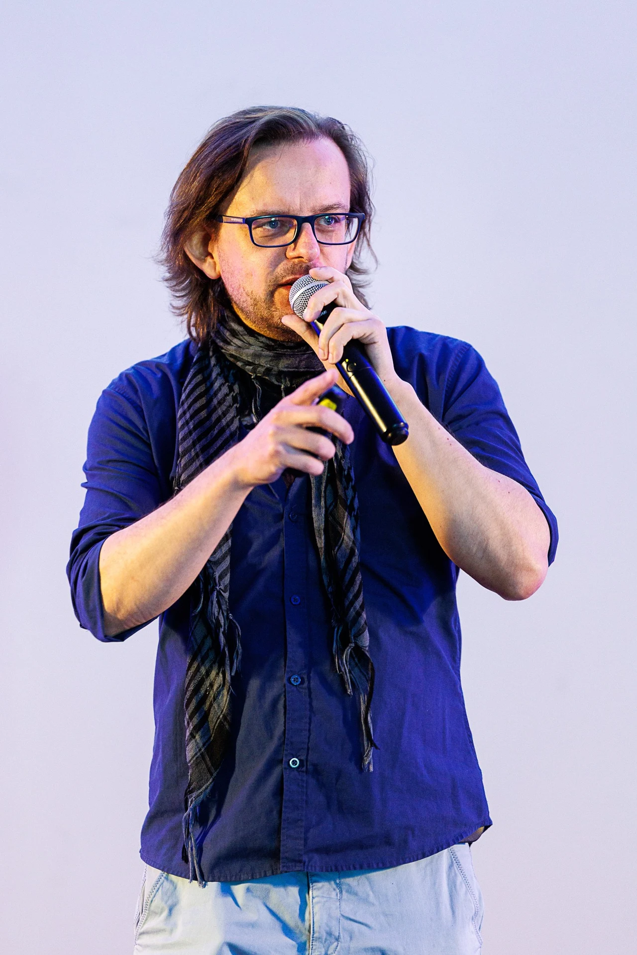 Artur Roguski Mężczyzna z długimi włosami i okularami trzyma mikrofon, ubrany w granatową koszulę i czarny szalik, wykonuje gesty rękami podczas wystąpienia na jasno oświetlonym tle.
