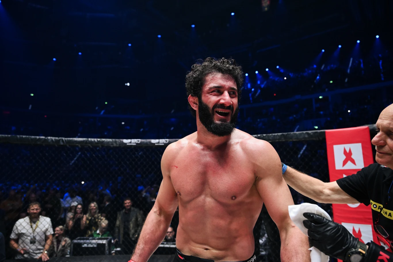 Mamed Chalidow - KSW mężczyzna z rozciętą wargą, muskularną sylwetką i uśmiechem na twarzy stoi na ringu MMA, otoczony przez ludzi w tle i asystenta dotykającego jego ramienia, mocne oświetlenie podkreśla atmosferę areny sportowej