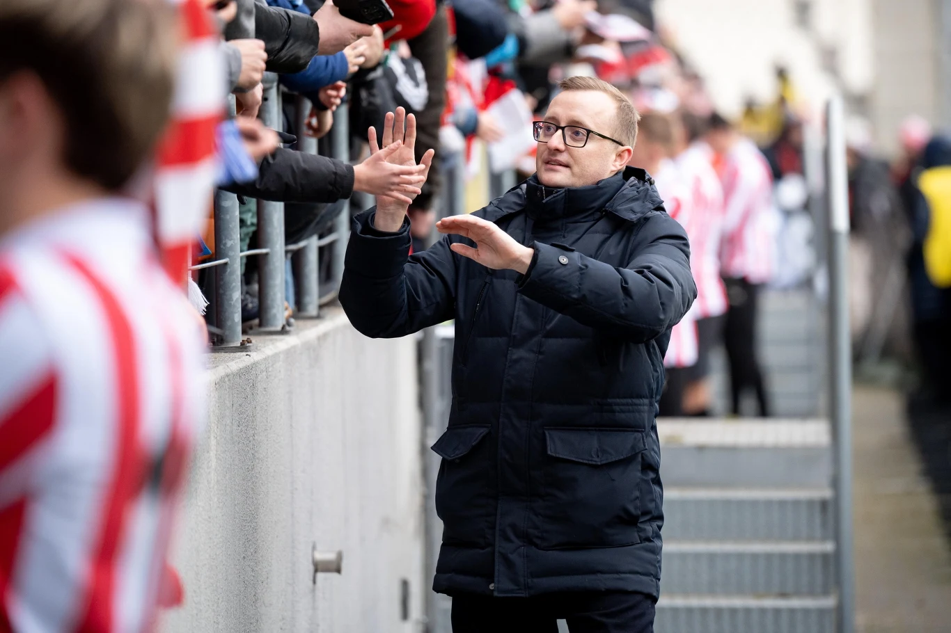 mężczyzna w okularach ubrany w ciemną kurtkę przybija piątki z kibicami w sportowych strojach za ogrodzeniem, atmosfera pełna entuzjazmu i wsparcia