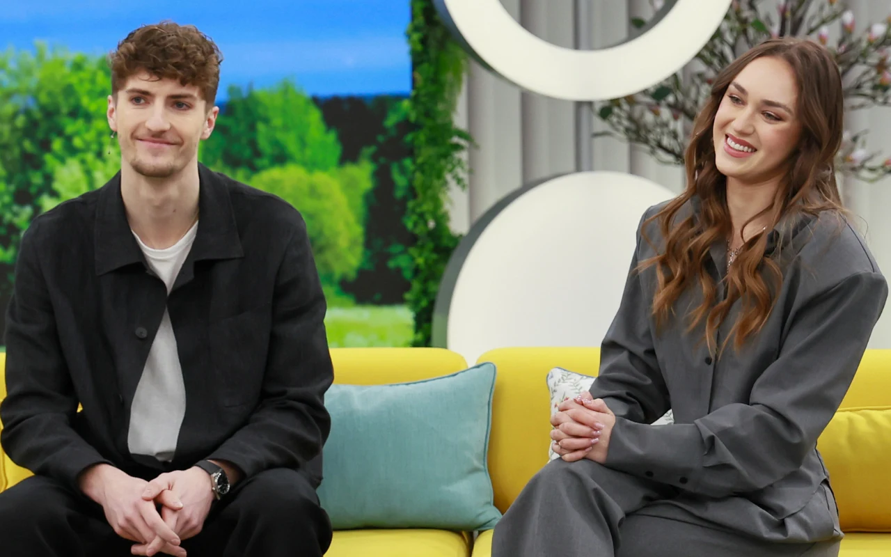 dwoje młodych ludzi siedzi obok siebie na żółtej kanapie, mężczyzna ubrany na czarno, a kobieta w szarym stroju, oboje uśmiechnięci, w tle nowoczesne wzory i zielone rośliny