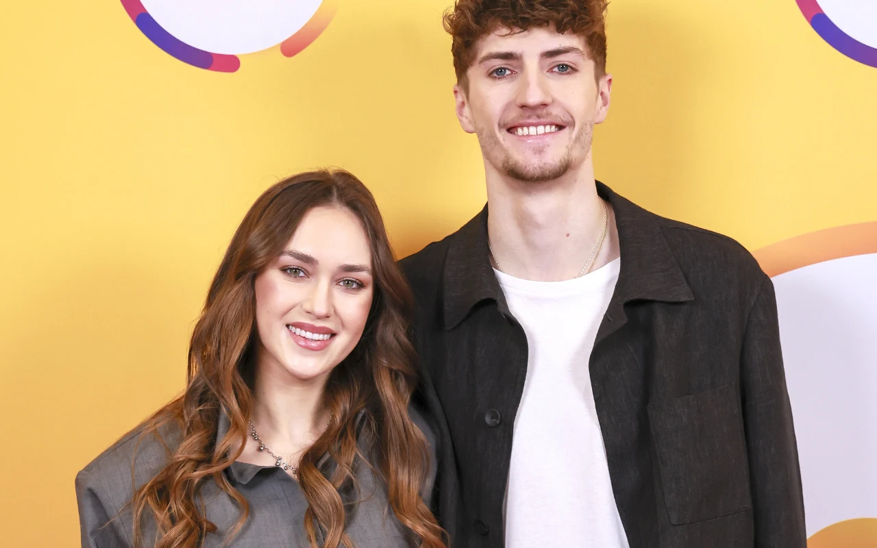 Młoda kobieta o długich, falowanych włosach i uśmiechnięty mężczyzna z kręconymi włosami stoją razem na tle żółtej ściany z kolorowymi, geometrycznymi elementami.